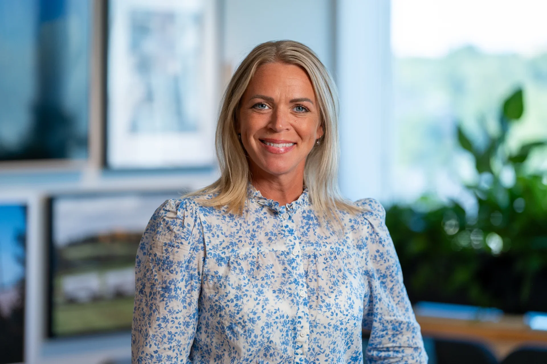 A smiling woman with blonde hair wearing a blue floral blouse in an office setting with a blurred background of windows and green plants.