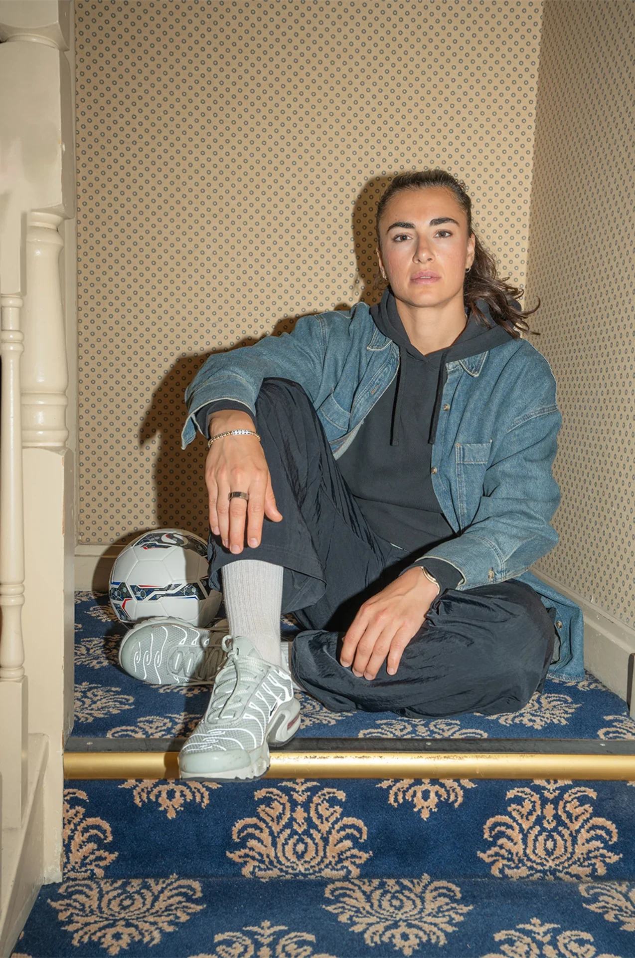 Young woman sitting on the floor in a corner with a soccer ball, wearing athletic clothing including sneakers, socks, and a denim jacket.