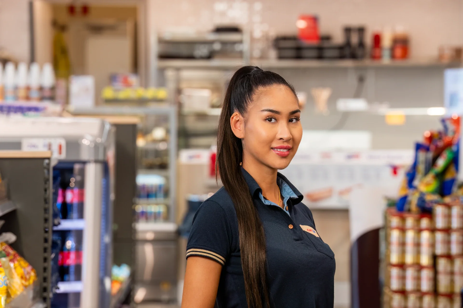 A store employee with long dark hair tied back, wearing a navy blue shirt with tan-striped cuffs and a name tag, stands in a grocery store aisle with shelves of snacks and condiments behind her.