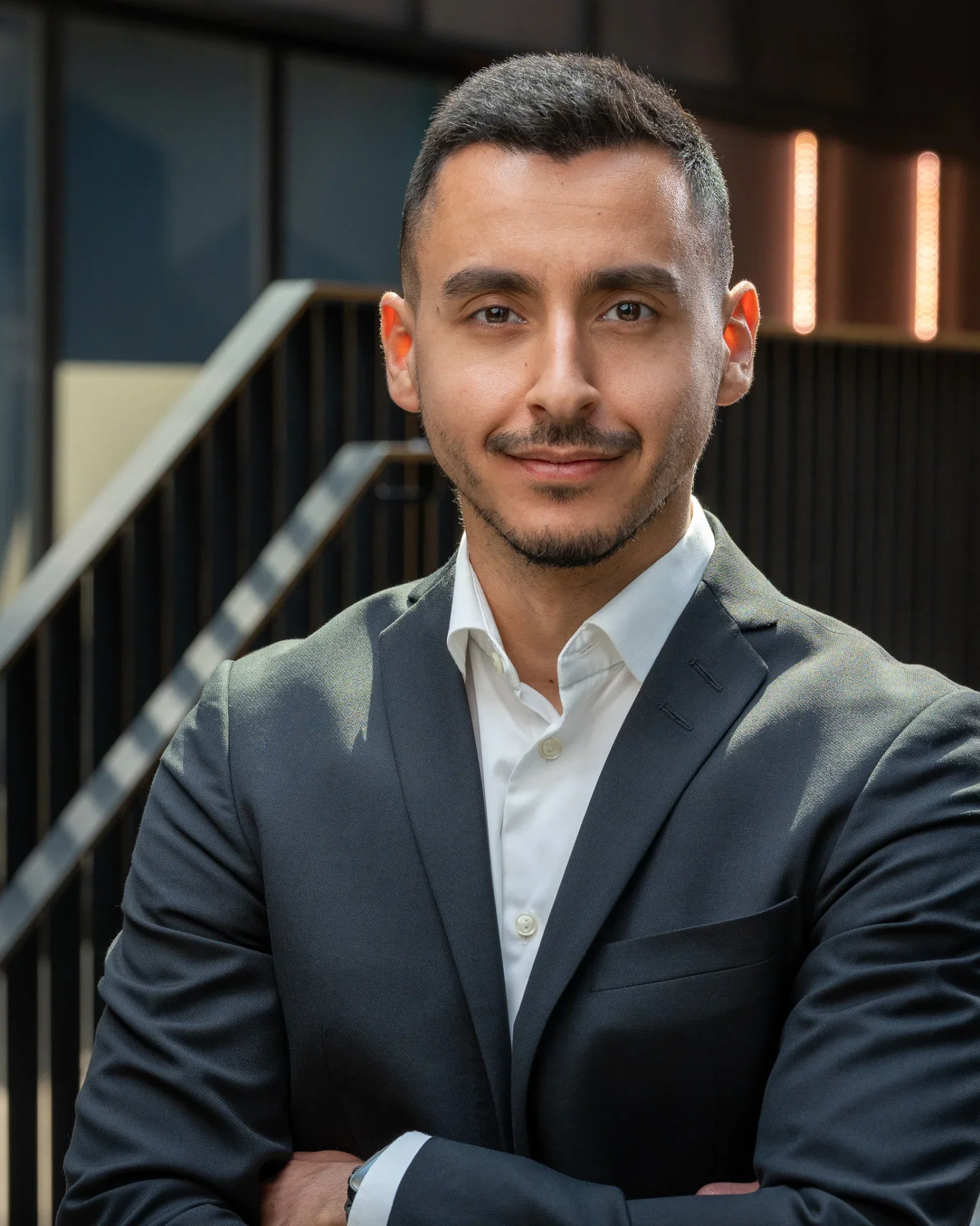 Close-up of a young man in a dark suit and white shirt, looking at the camera with a confident expression, standing in front of a modern office background.