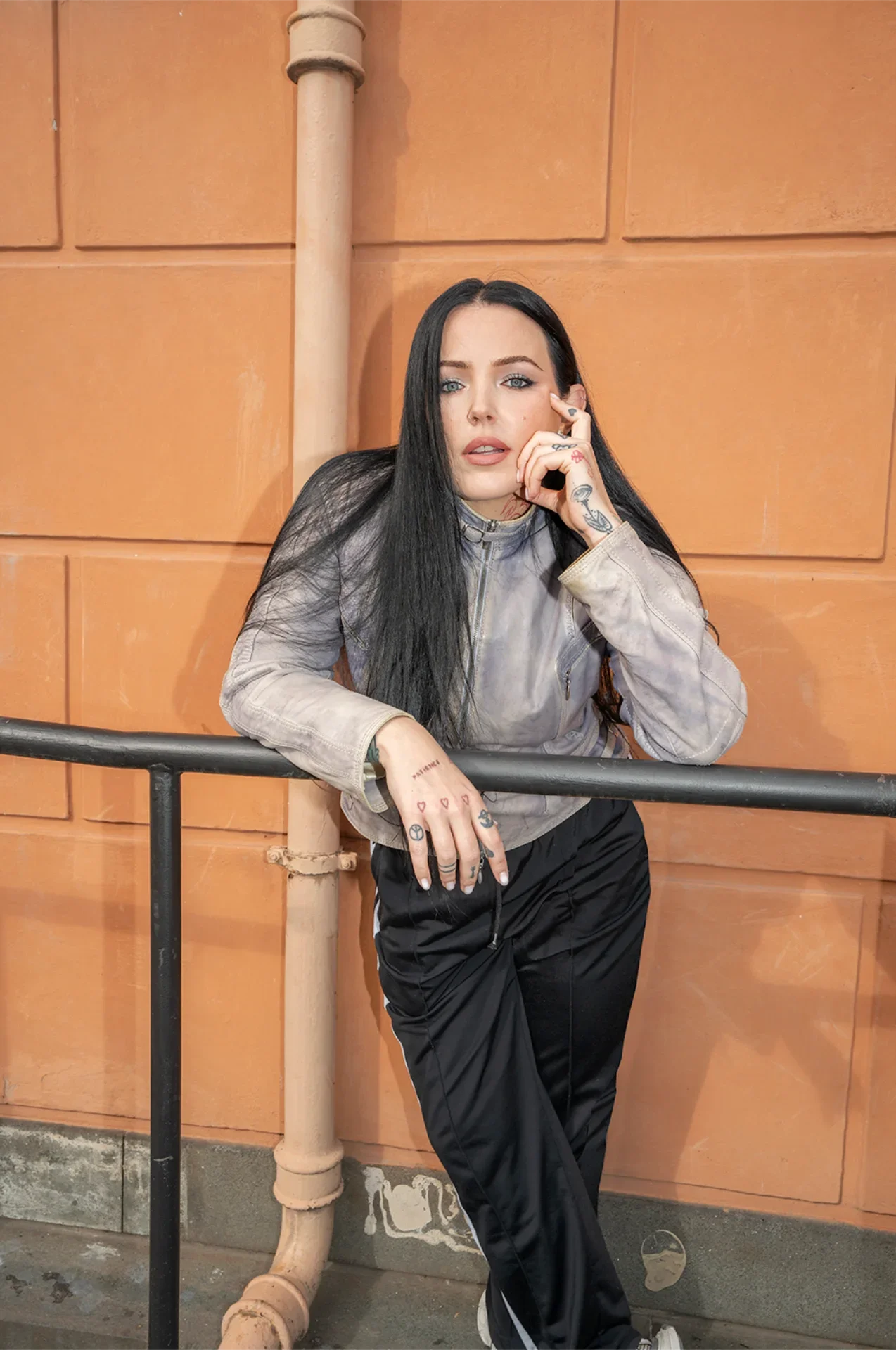 A young woman with long black hair, tattoos, and piercing blue eyes leaning against a metal railing in front of an orange wall, wearing a gray jacket and black pants.