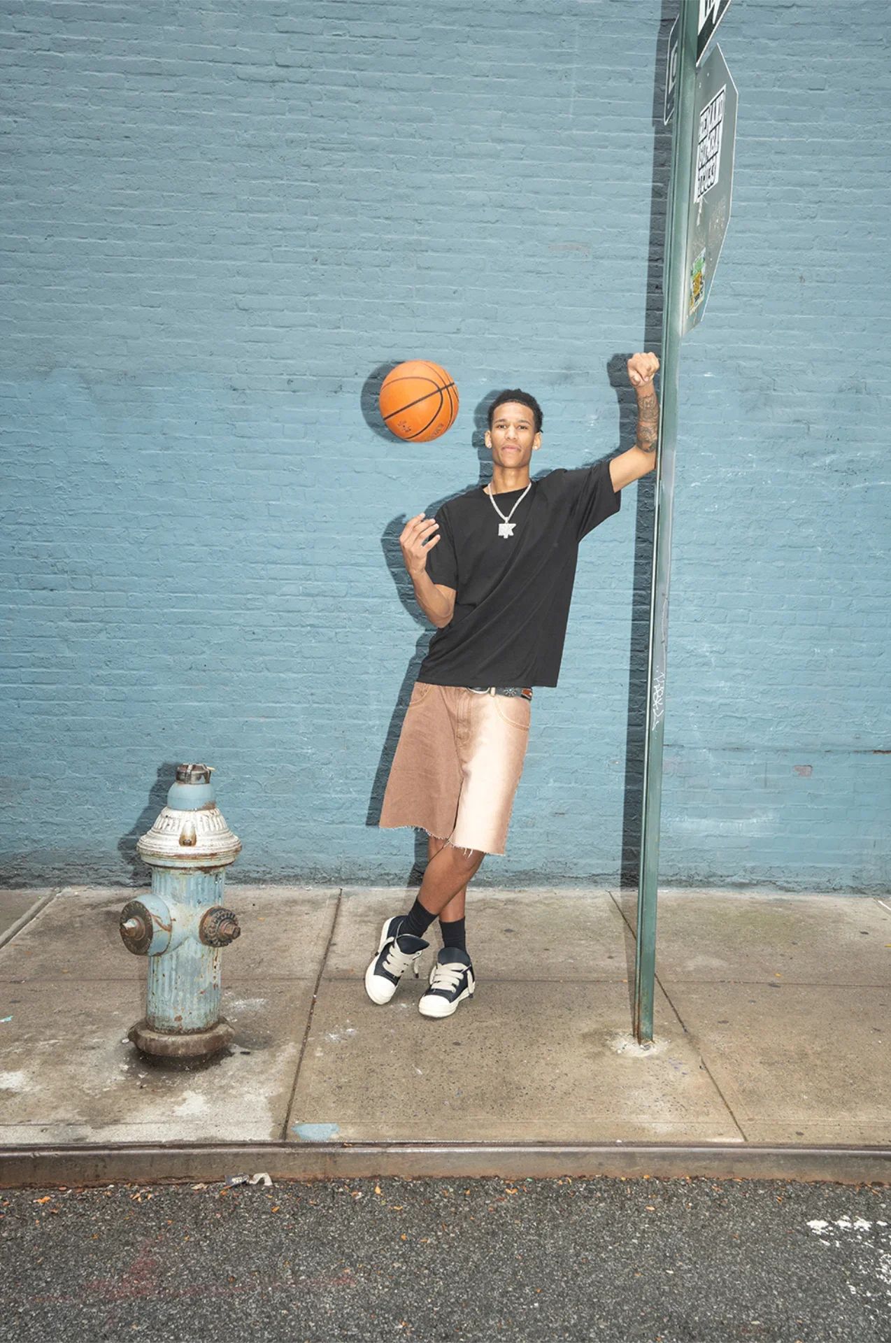 A young man standing against a blue brick wall, holding a basketball in midair and flexing his right arm, wearing a black t-shirt, beige shorts, black socks, and white sneakers with black accents.