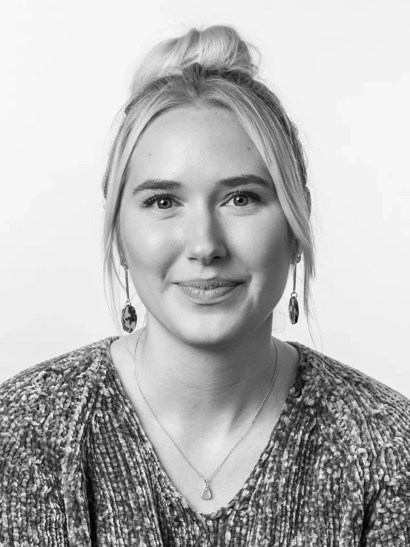 Black and white portrait of a woman with light-colored hair styled in an updo, wearing a textured sweater, earrings, and a necklace.