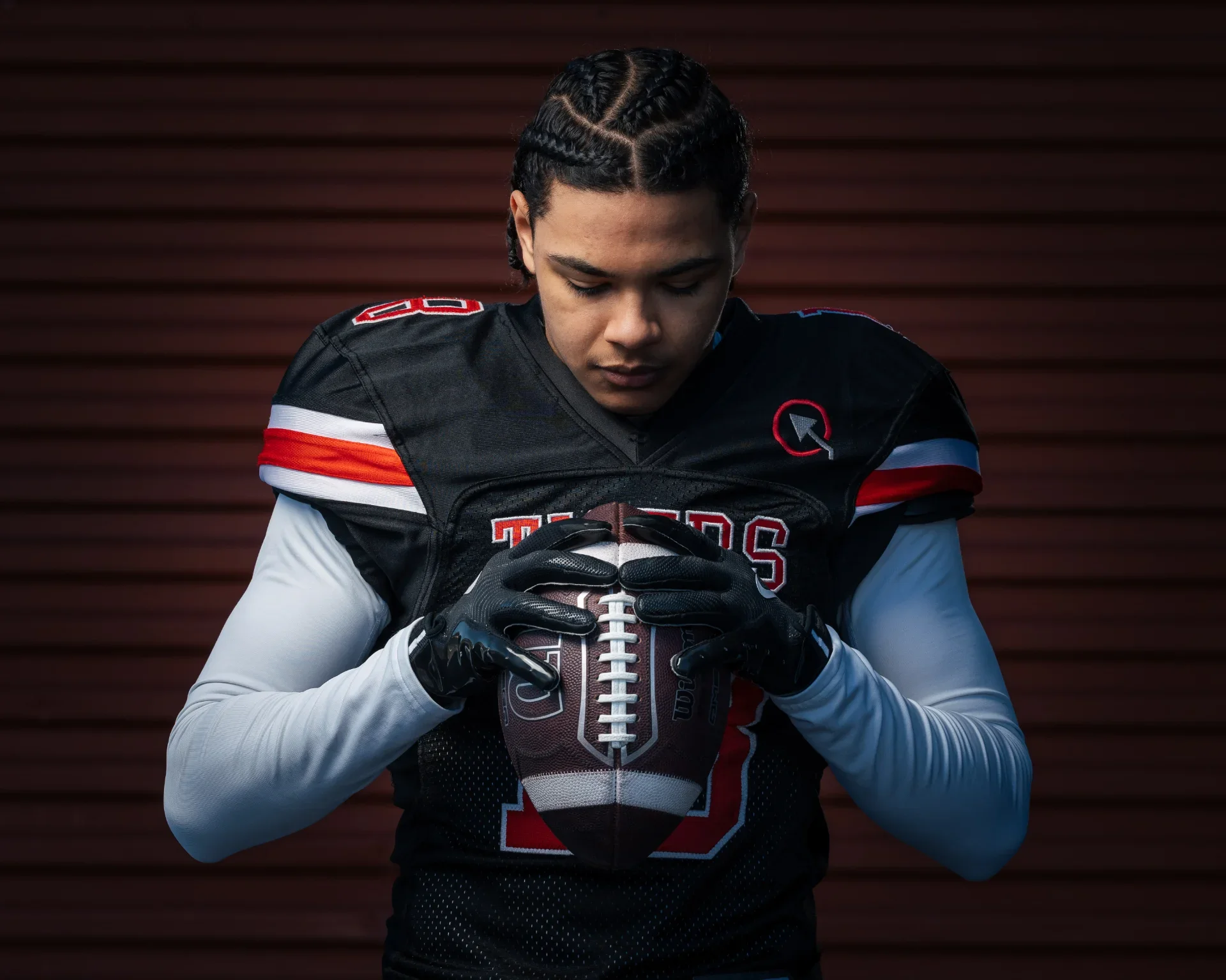 Young football player in a black and red uniform holding a football with both hands, looking down at it against a brown wooden background.