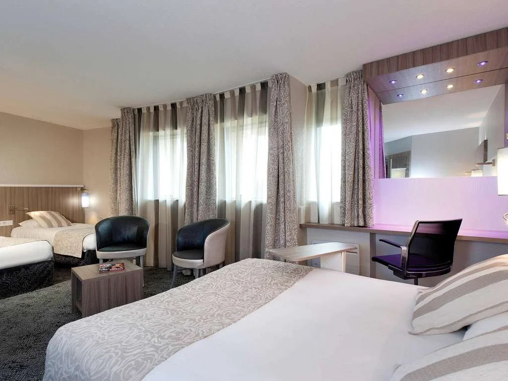 Chambre d'hôtel avec deux lits, fauteuils, bureau, et grandes fenêtres avec rideaux.