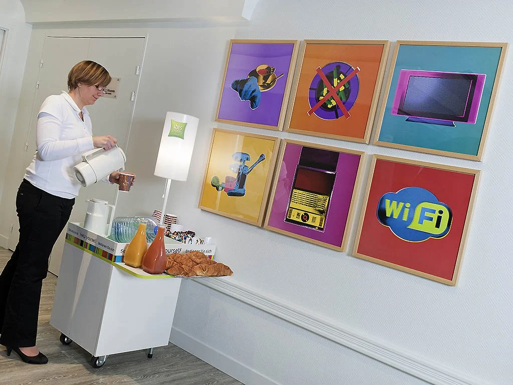 Une femme se sert une boisson dans un espace qui semble être une salle de pause, avec un petit buffet. Sur le mur à droite, il y a six cadres avec des illustrations ou icônes, notamment une télévision, une icône Wi-Fi, et d'autres symboles liés à la technologie ou aux appareils électroniques.