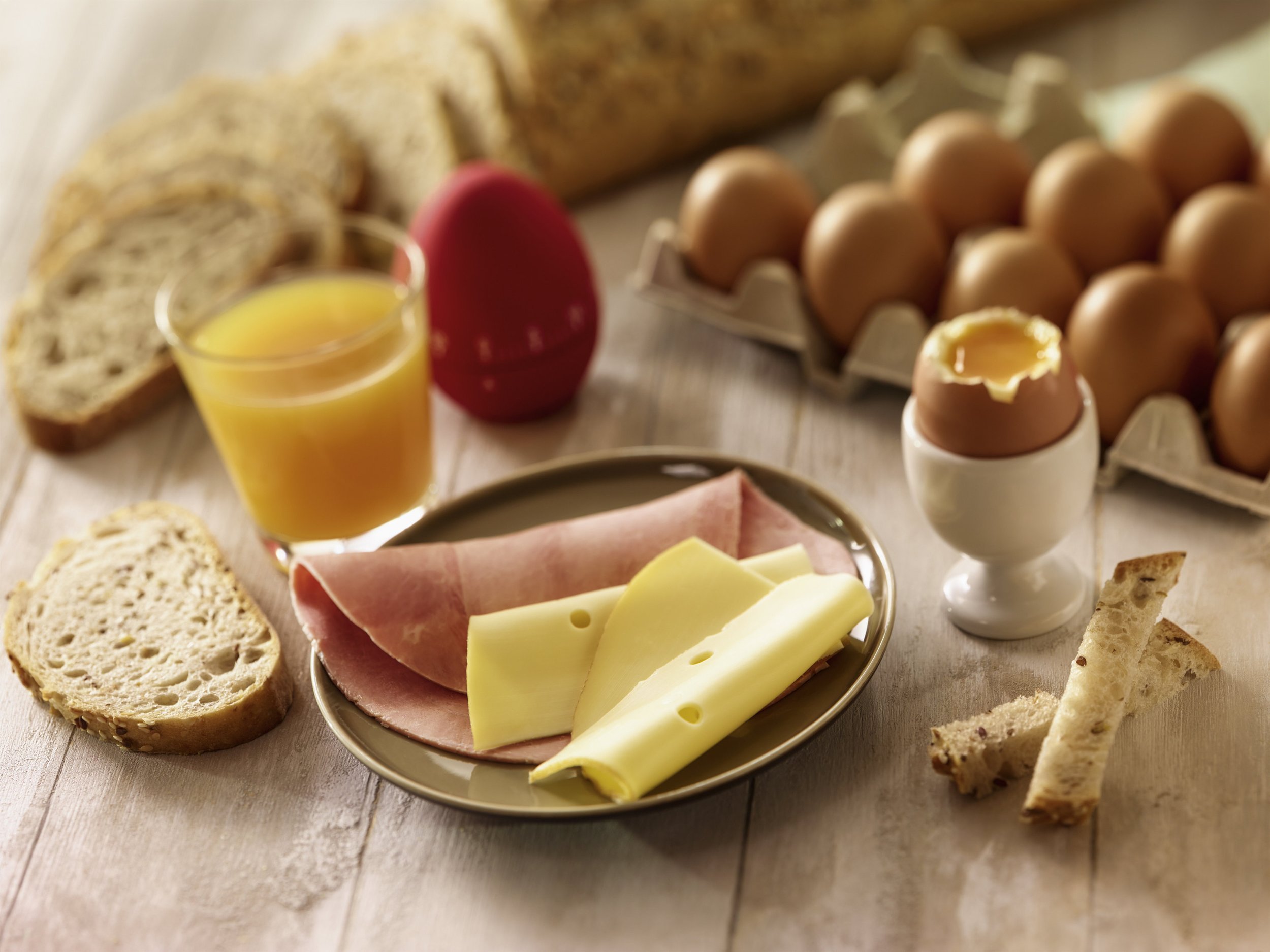 A breakfast spread with sliced bread, a glass of orange juice, a red soft-boiled egg, a dozen eggs in a carton, sliced ham and cheese on a plate, and breadsticks on a wooden surface.