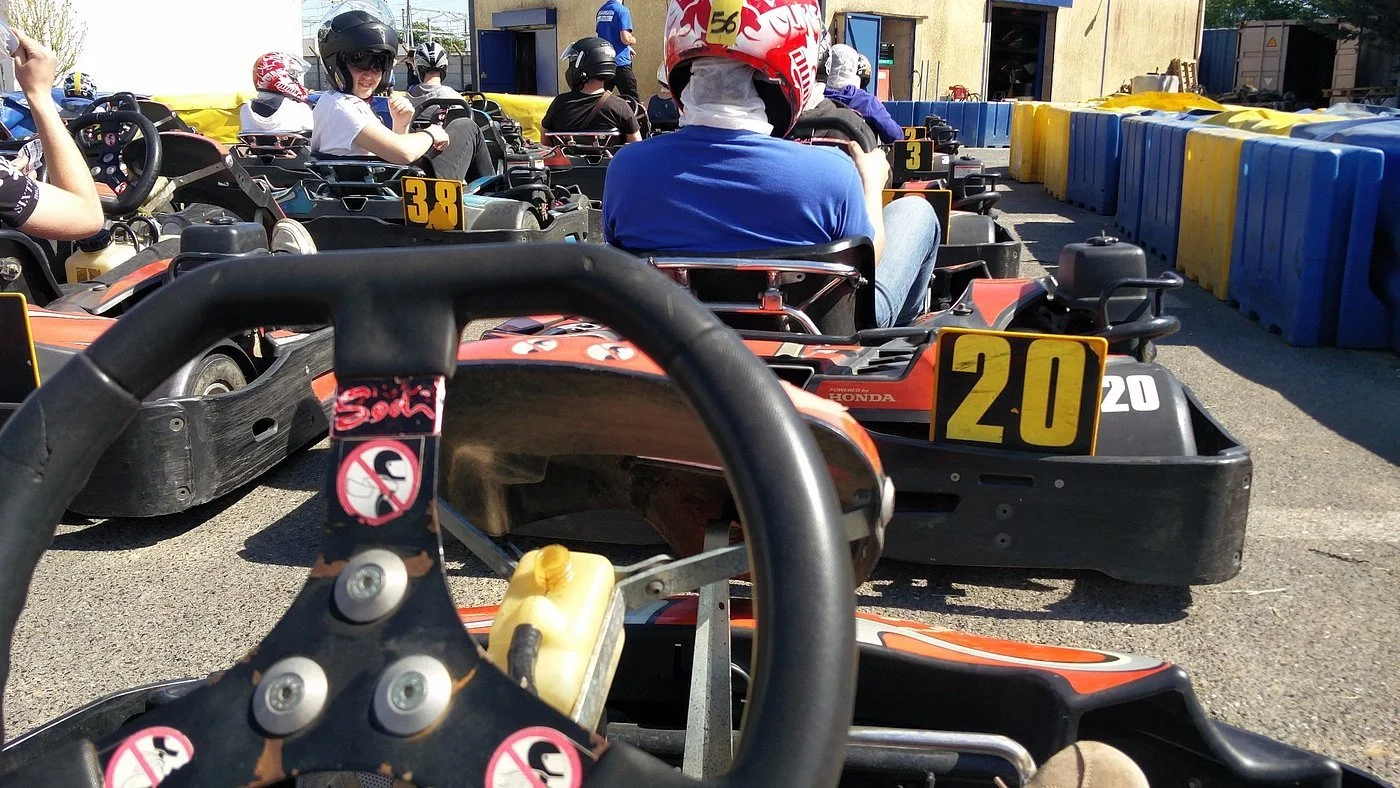 L'image montre un groupe de personnes en karts de course, assises dans leurs véhicules, équipées de casques. La photo est prise depuis l'intérieur d'un kart, montrant le volant, avec d'autres karts alignés en ligne devant eux, sur un circuit ou une zone de course avec des barrières en plastique bleu et jaune.