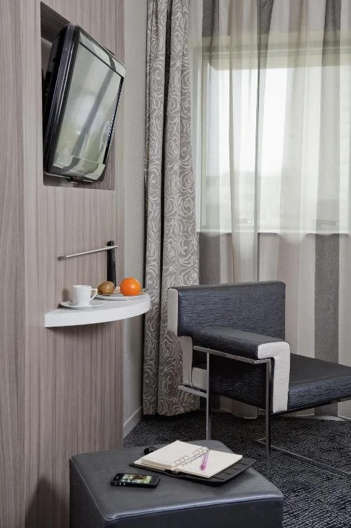 Une chambre d'hôtel avec un mur en bois, une télévision à écran plat fixée au mur, une petite étagère blanche avec une tasse, un croissant, une orange et un autre fruit, une chaise noire et blanche, une fenêtre avec des rideaux légers, et un coffre noir avec un cahier, un stylo et un téléphone portable dessus.