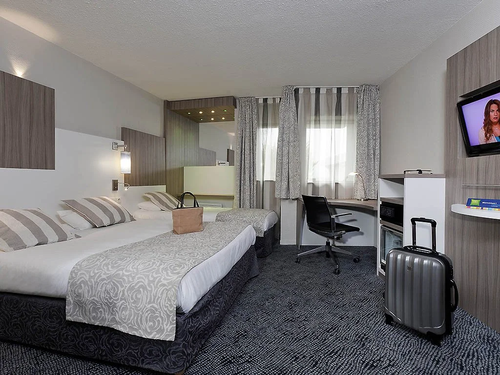 Hotelzimmer mit Doppelbett, Schreibtisch, an der Wand montiertem Fernseher, Rollkoffer und Fenster mit Vorhängen.