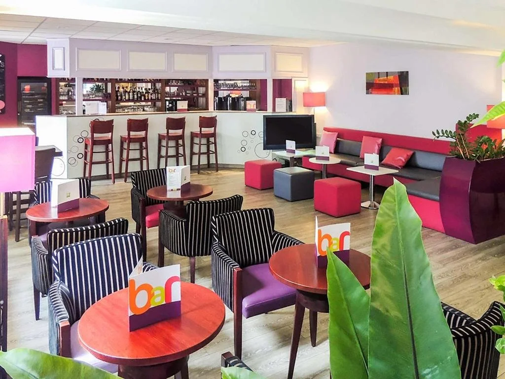 Un café ou bar moderne avec des tables rondes et des chaises à rayures, un comptoir avec des tabourets rouges, un coin salon avec un mur gris et une banquette rose, un téléviseur, des plantes vertes et une décoration colorée.