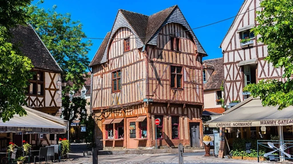 Une maison à colombages avec un toit en pente, entourée d'autres maisons à colombages dans une rue pavée, avec des arbres verts et la lumière du soleil.