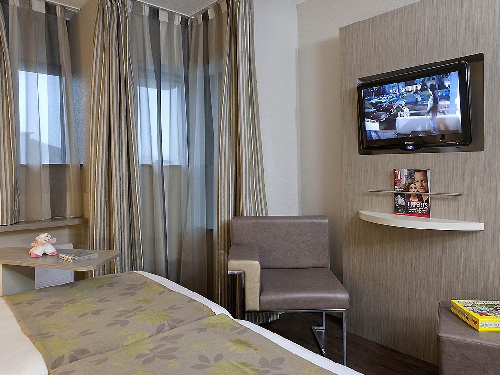 Chambre d'hôtel avec lit, télévision à écran plat, fauteuil, fenêtre avec rideaux, table de chevet avec jouet et livres, mur coloré et décoratif.