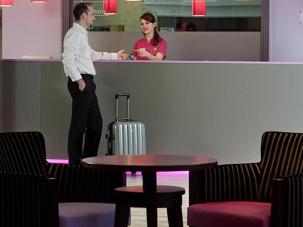 Un homme et une femme souriants échangent à un comptoir d'hôtel ou d'aéroport, avec une valise à roulettes à côté d'eux, dans un espace d'attente moderne.