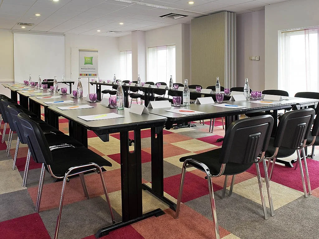 Salle de réunion avec tables en forme de U, chaises noires, bouteilles d'eau, verres en plastique violet, et papiers avec stylos.
