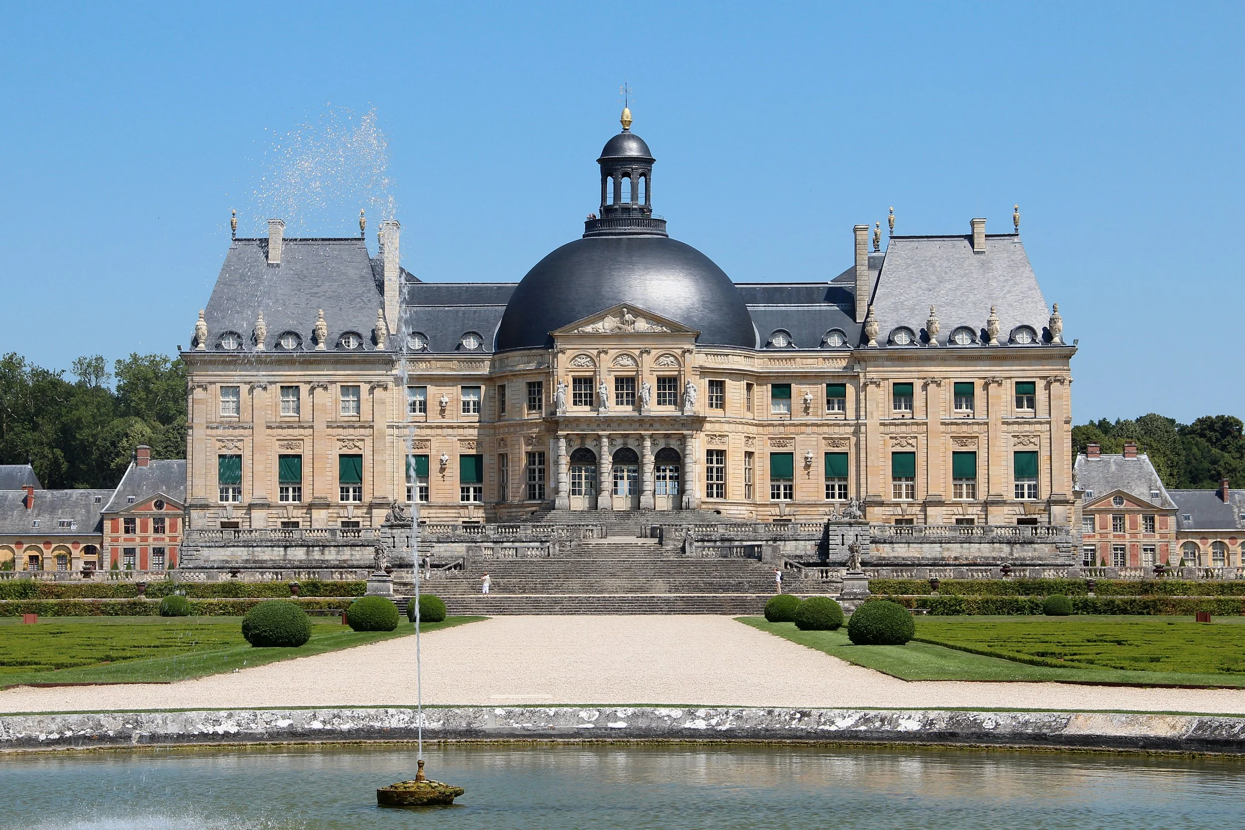 Un château élégant avec un grand dôme noir, plusieurs statues ornant la façade, des fenêtres avec volets verts, et un escalier menant à l'entrée principale, entouré de jardins bien entretenus et d'une fontaine au premier plan.