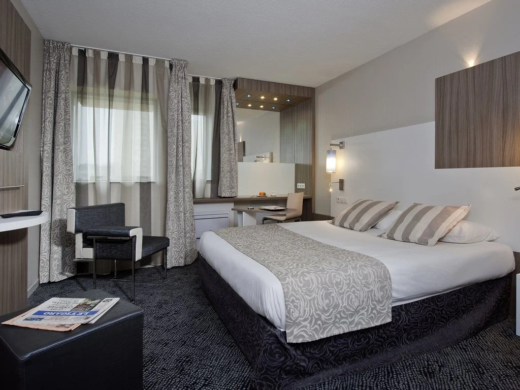 Chambre d'hôtel avec lit double, fauteuil, bureau, télévision murale, et fenêtres avec rideaux