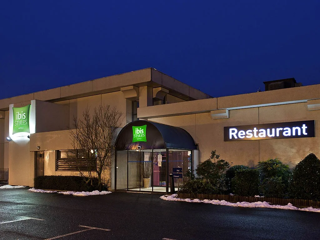 Entrée d'un hôtel Ibis Styles avec un restaurant à côté, il fait nuit, il y a des arbustes et de la neige sur le sol.