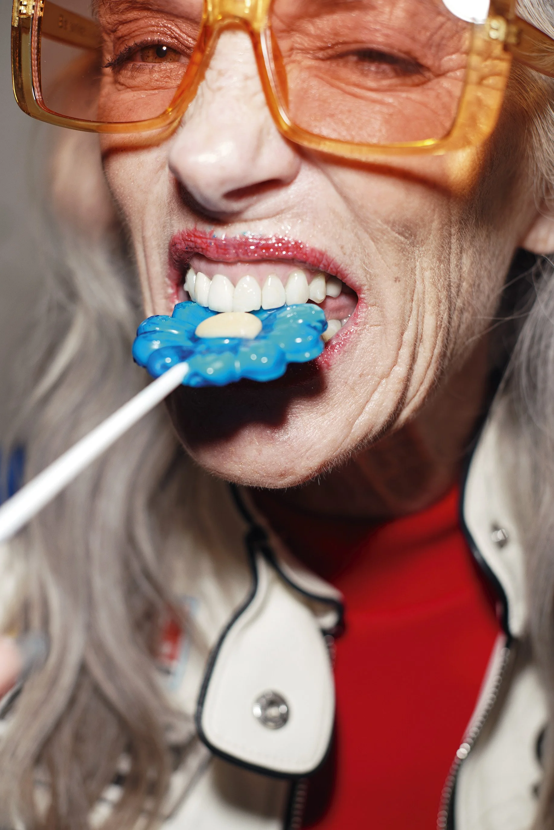 Une femme avec des lunettes orange et un blazer blanc, mordant dans une sucette bleue en forme de fleur.