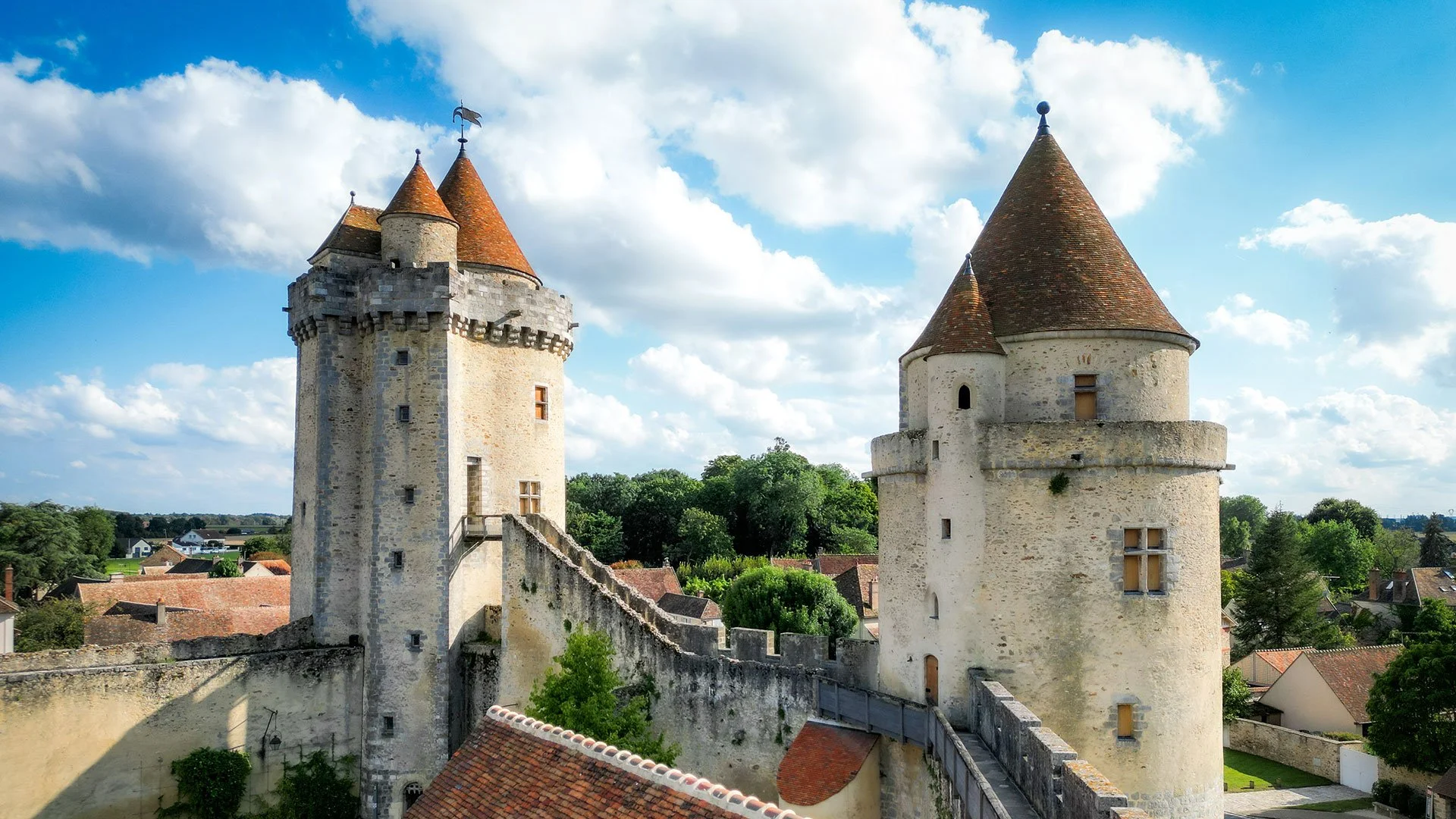 Deux tours de château avec des toits en forme de cône en tuile orange, entourées de murs en pierre, avec un ciel bleu et des nuages blancs, dans un paysage verdoyant.