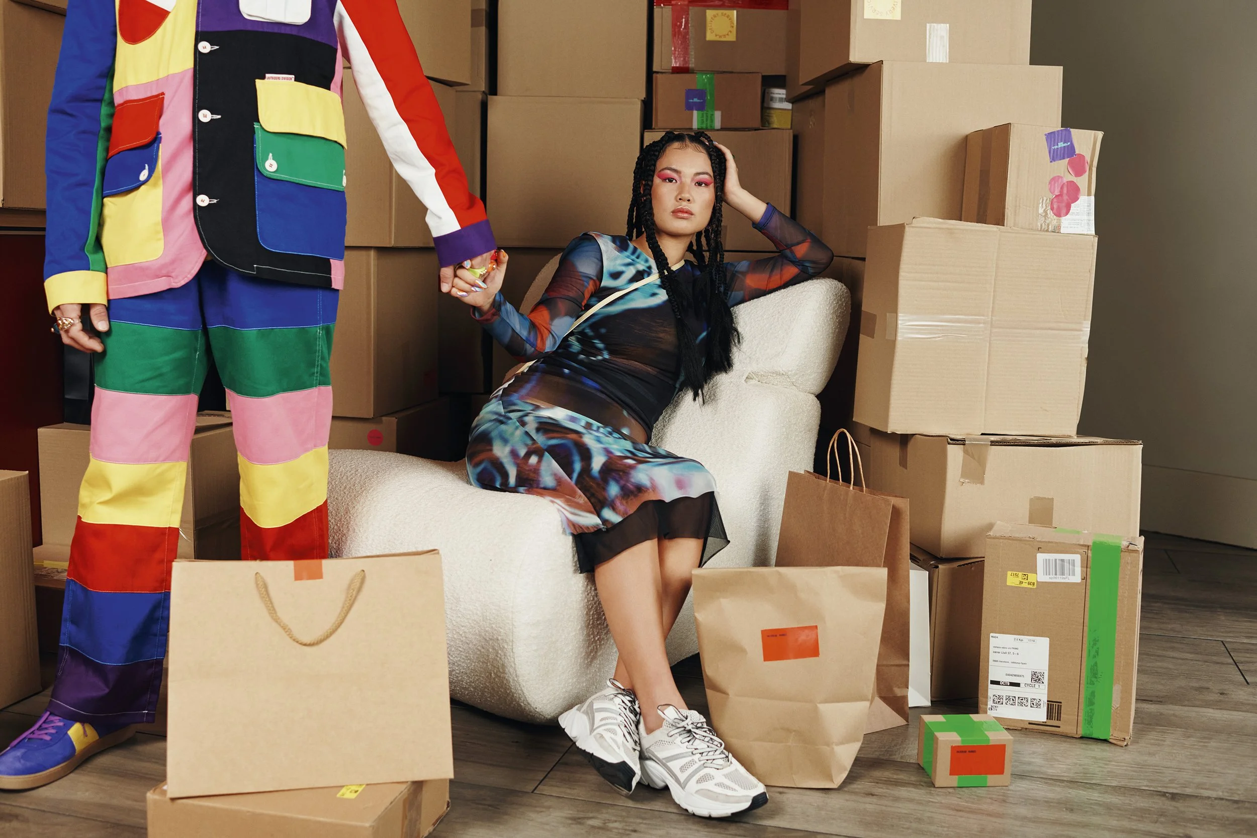 Une femme assise parmi des cartons de déménagement, tenant la main d'une autre personne en costume coloré, dans une pièce remplie de boîtes et de sacs en papier.