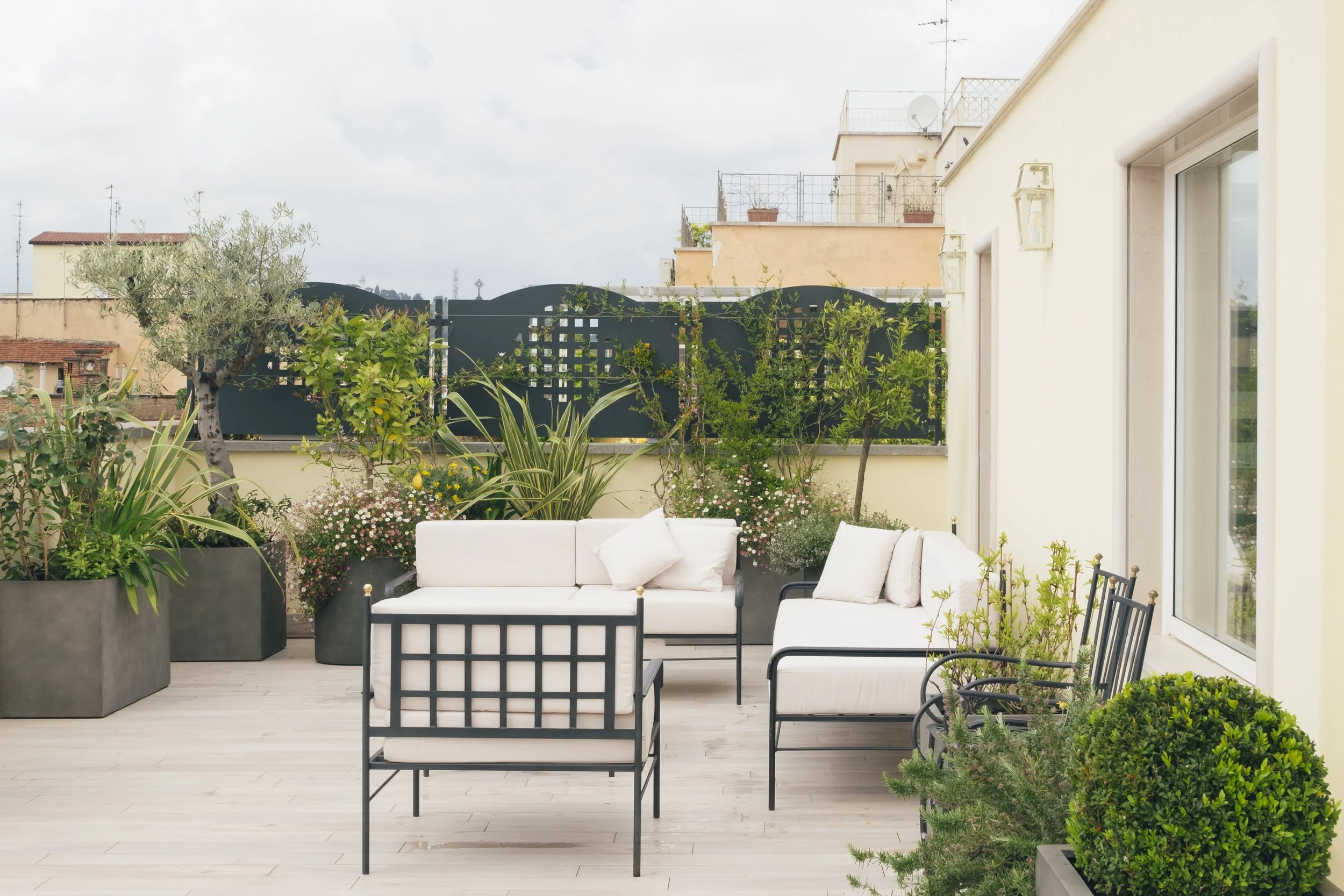 Balcone con sedie e divani bianchi, piante e fiori, sullo sfondo edifici e tettoie di una città.