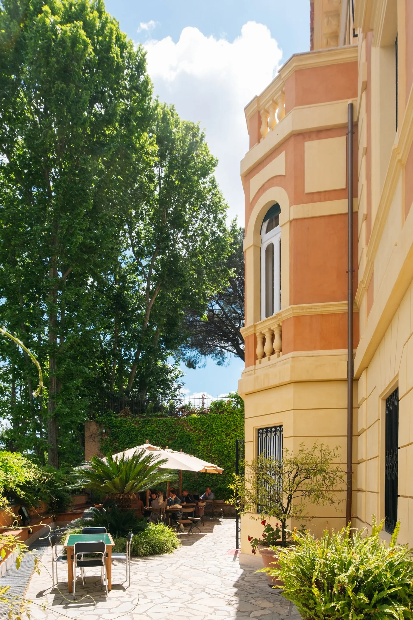 Un cortile con tavoli e sedie occupati da persone, piante verdi e un grande albero, con un edificio di colore giallo e rosa con balconi e finestre, e un cielo soleggiato con alcune nuvole.