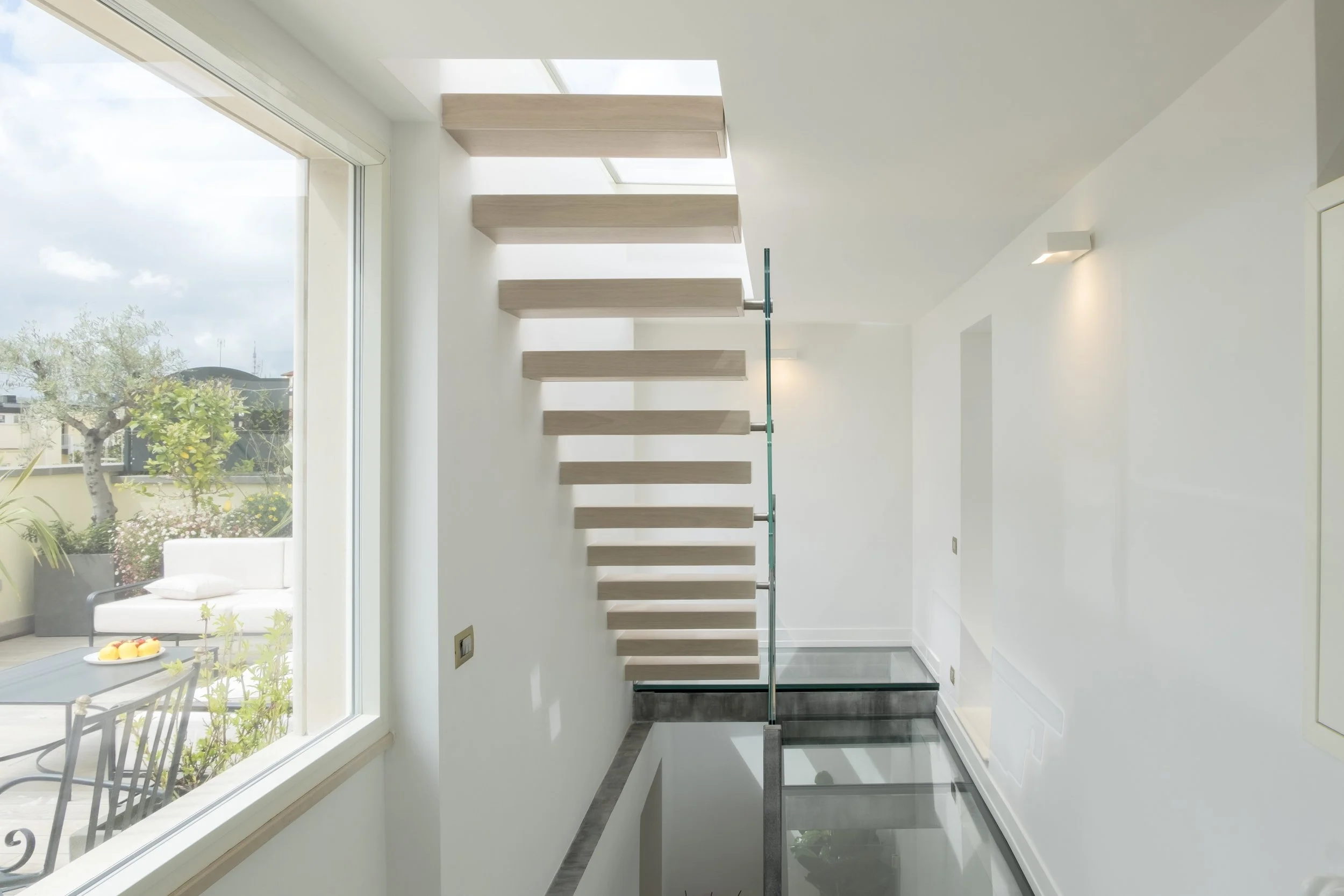 Interno moderno con designer scala a sbalzo in legno e parapetto in vetro, pareti bianche, finestra grande che mostra un giardino esterno con piante e sedie.