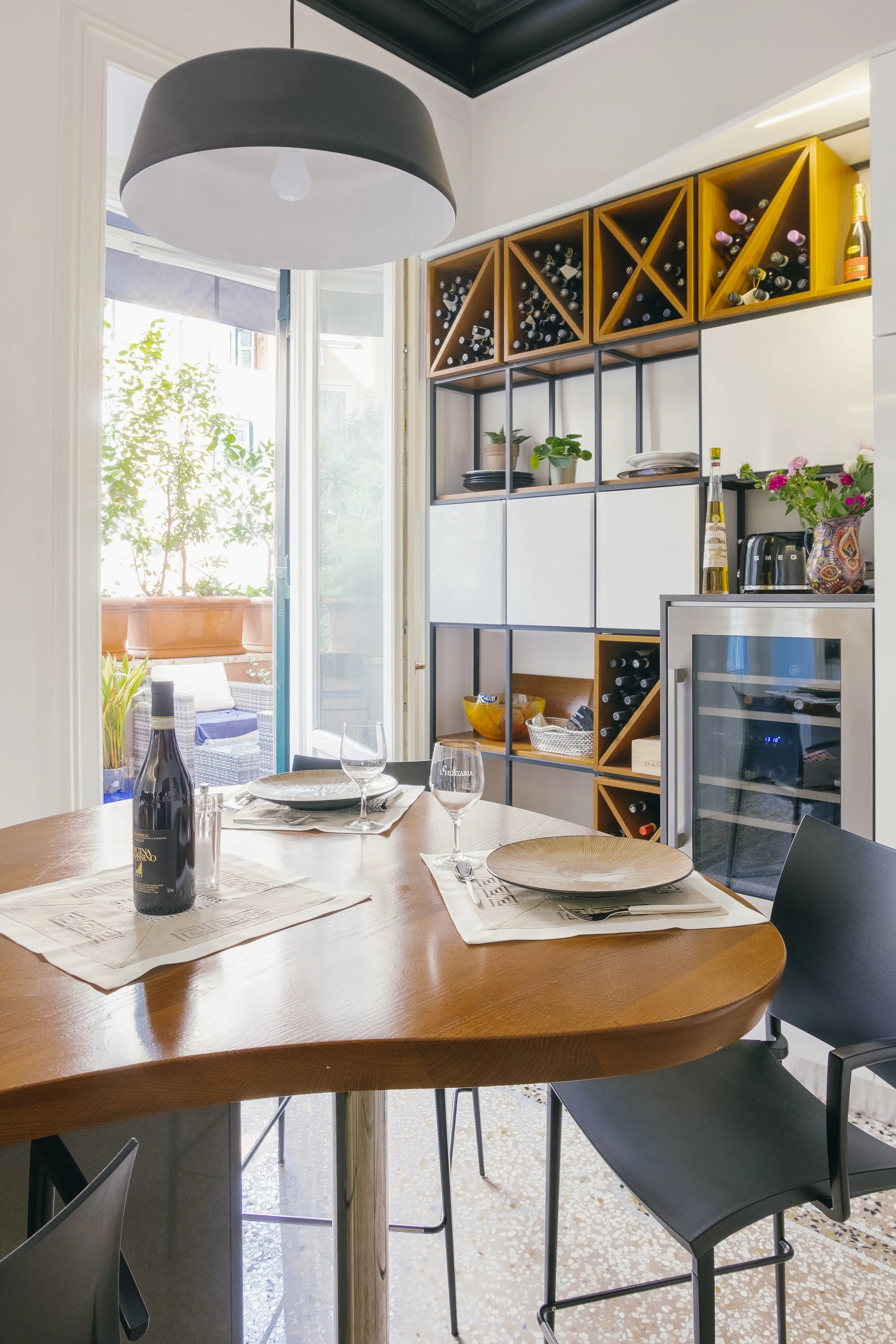 Interno di una cucina con tavolo da pranzo, bicchieri, bottiglie di vino, piatti e posate, e una parete con scaffali di vino e piante. Apertura su terrazza con piante e seduta.