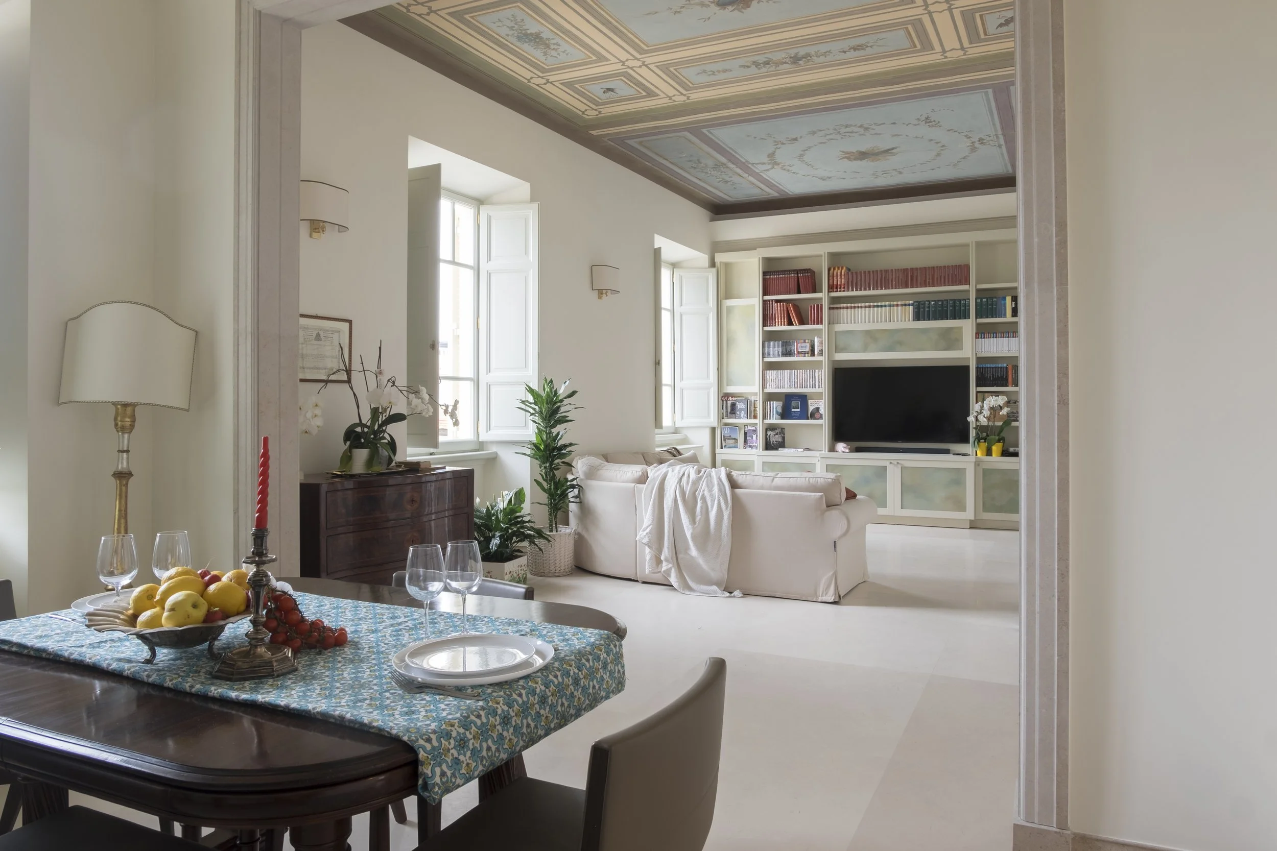 Interior di una stanza luminosa con soffitto decorato, pareti bianche, e mobili eleganti. In primo piano, tavolo con frutta e bicchieri, sullo sfondo divano, libreria con libri e televisore, molte piante e finestre aperte.