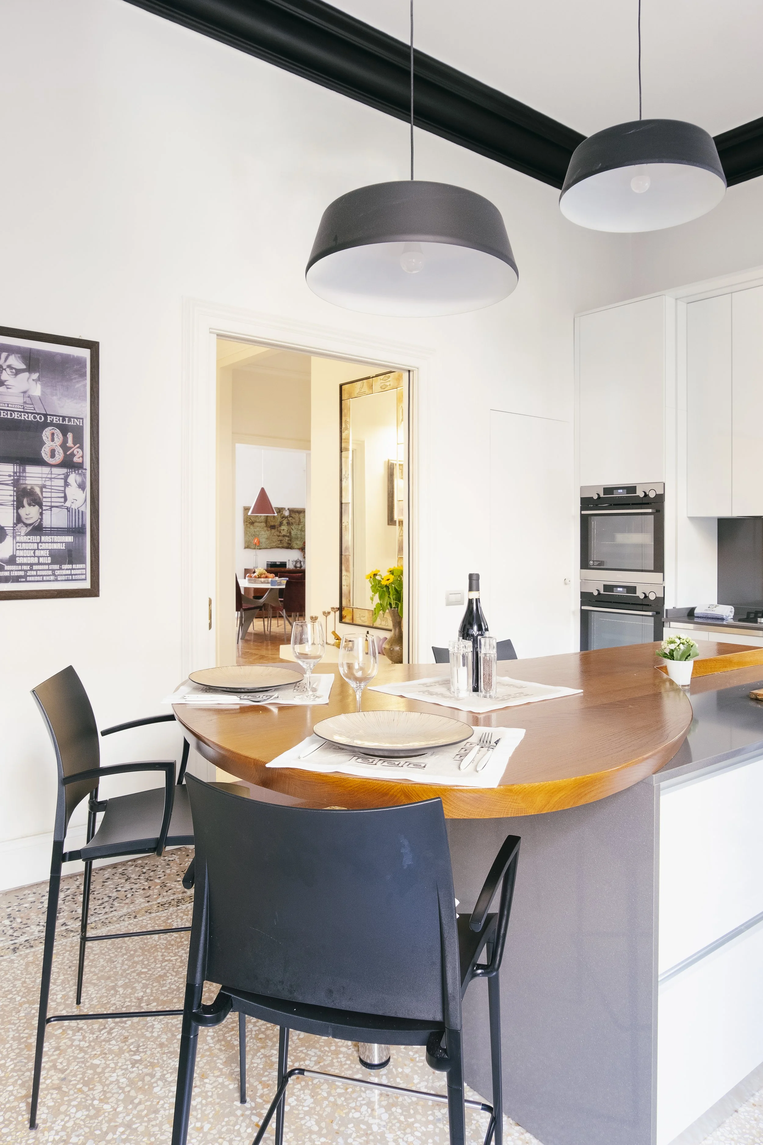Interno di una cucina moderna con tavolo rotondo in legno, sedie nere, illuminazione a soffitto di due lampade nere, pareti bianche, armadietti bianchi, forno incorporato, piccoli fiori e bottiglia di vino sul tavolo.