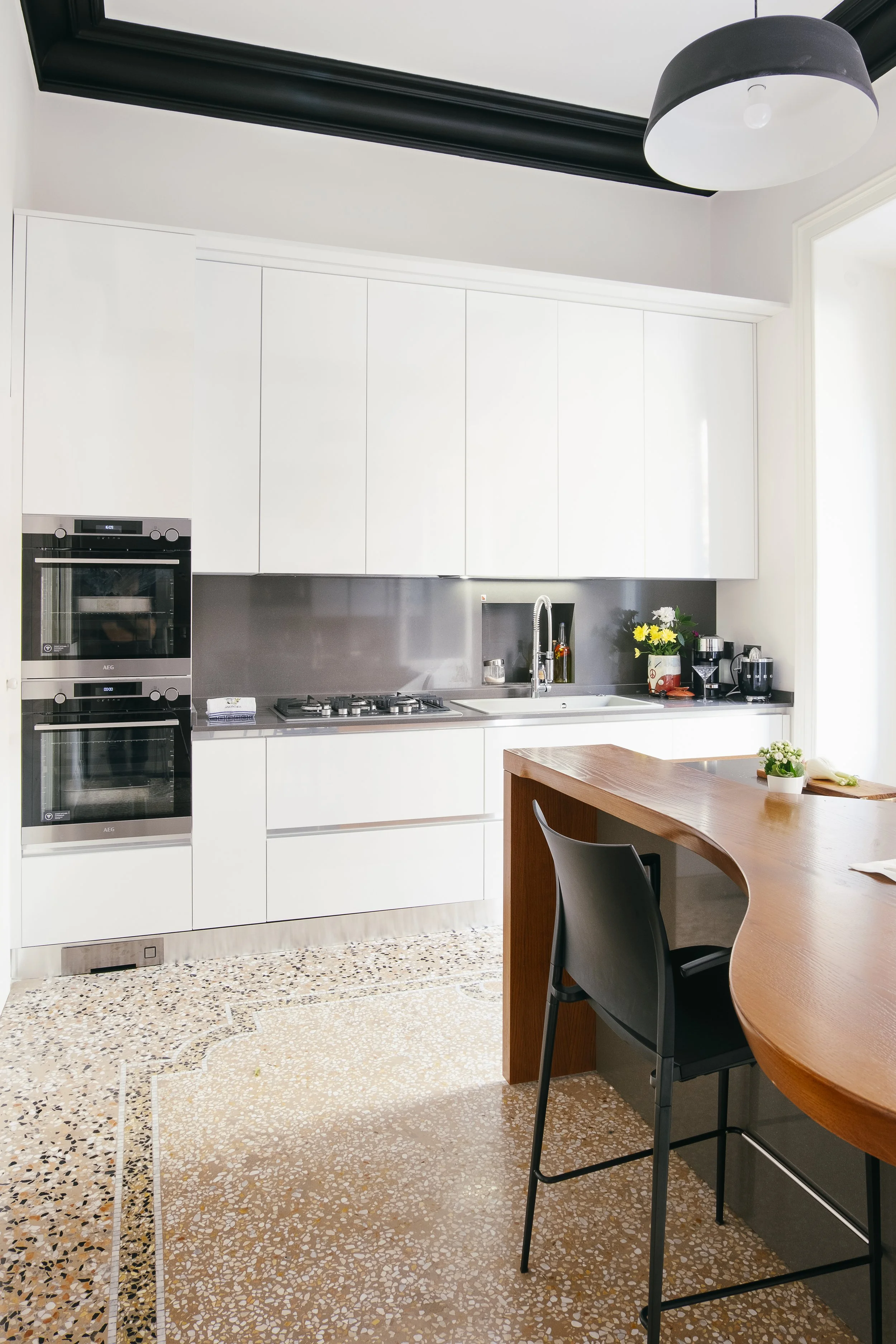 Cucina moderna con pareti bianche, armadi a muro, piano cottura a quattro fuochi, lavello con rubinetto, forno a microonde, alcuni oggetti da cucina e un tavolo di legno con una sedia nera.
