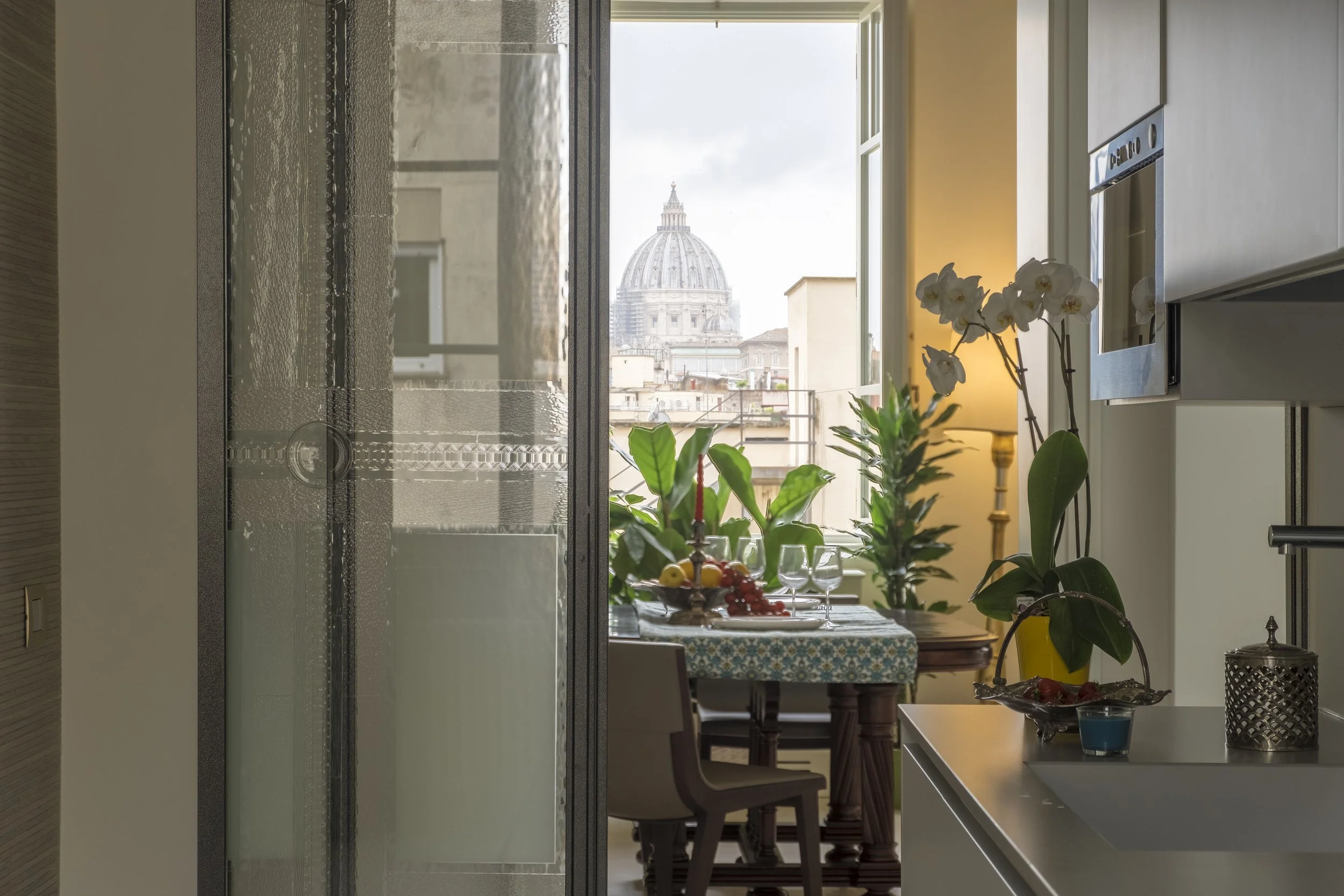 Vista di un'ampia finestra che mostra una cupola di una chiesa nel centro di Roma, con arredi e piante in primo piano all'interno di una cucina o sala da pranzo.
