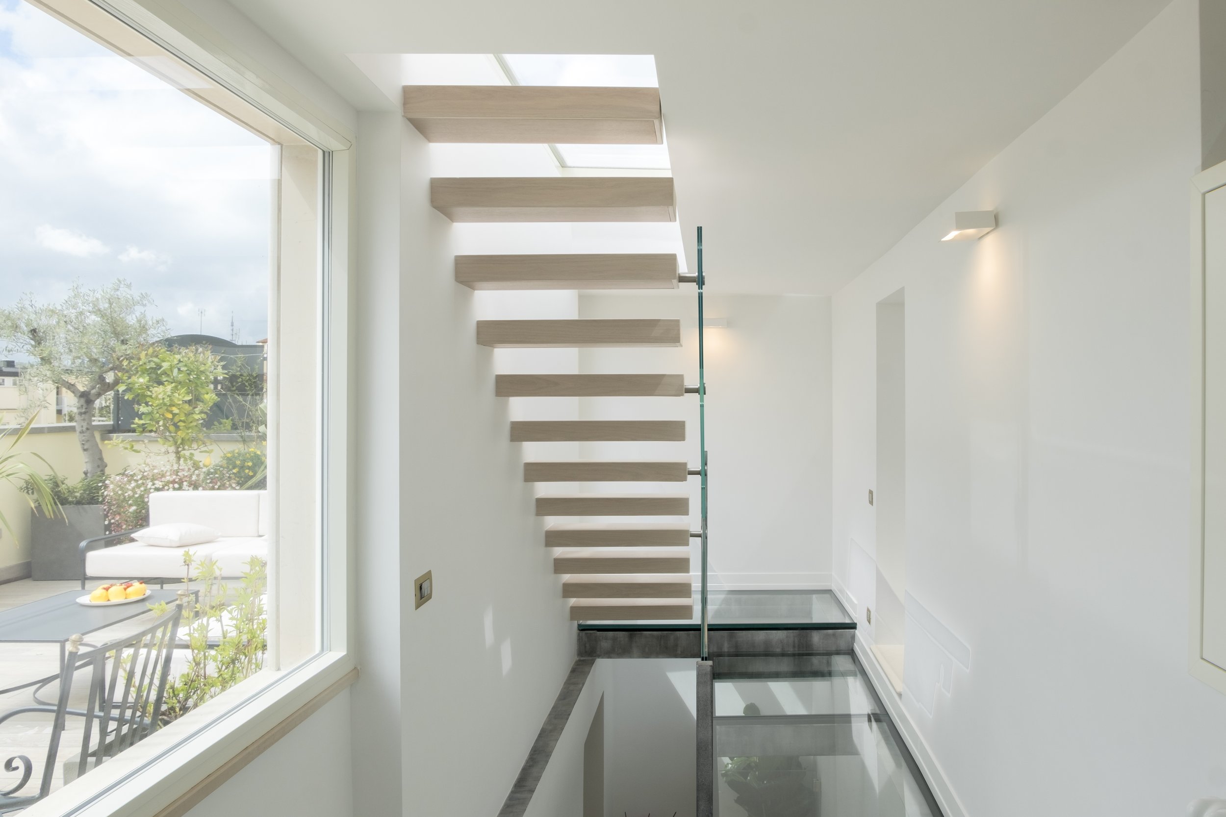 Interne con scale a giorno in legno e vetro, pareti bianche, grande finestra che lascia entrare molta luce naturale, vista esterna di un giardino con piante e terrazza con tavolo e sedie.