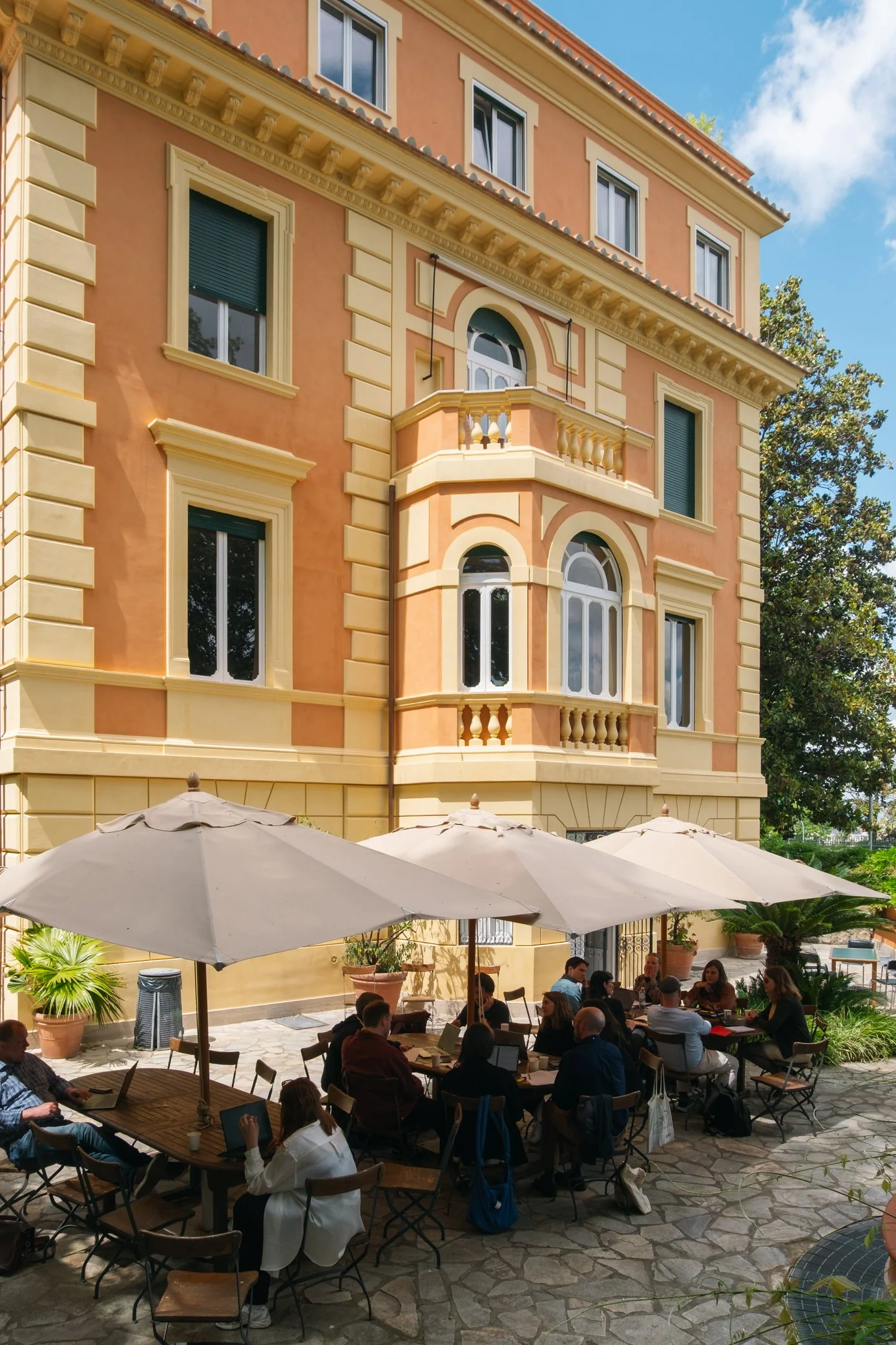Cortile con tavoli e ombrelloni, persone sedute che lavorano o socializzano, edificio storico sullo sfondo, cielo soleggiato e alberi.