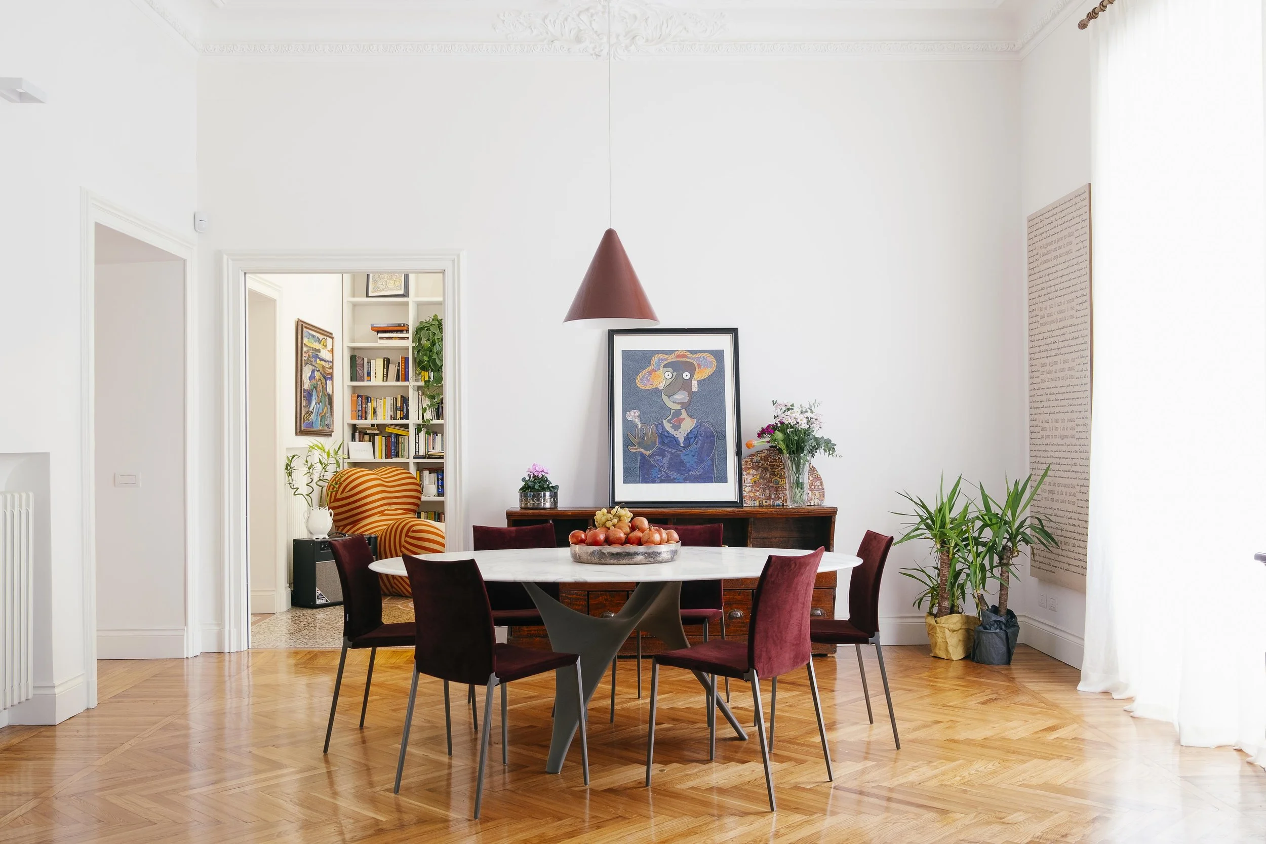 Sala da pranzo con tavolo rotondo e sedie rosse, pareti bianche decorate, quadri e piante, con soffitto dettagliato e lampadario appeso.