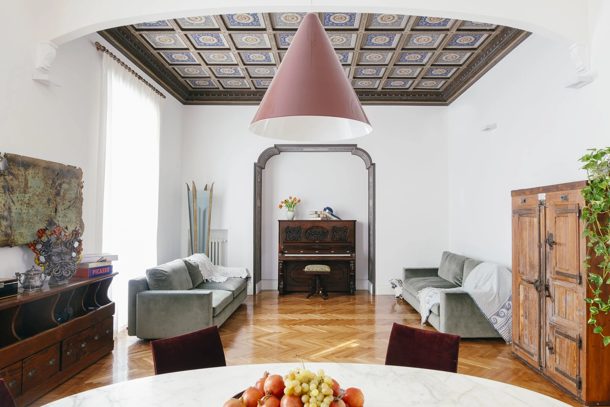 Interno di una stanza con soffitto decorato, due divani, un pianoforte e vari segreti d'antiquariato, con una finestra luminosa con tende bianche.