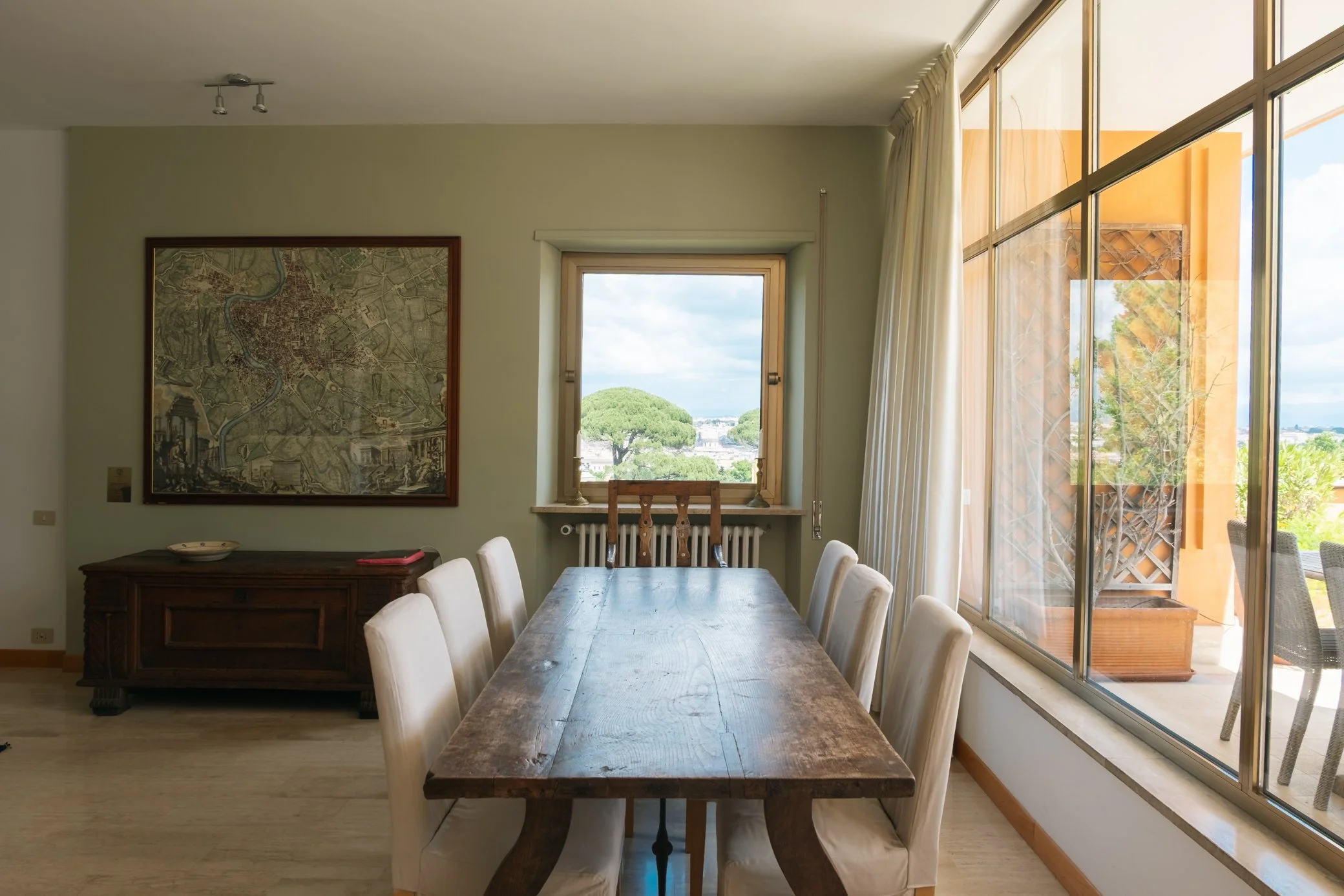 Sala da pranzo con una lunga tavola di legno, sedie imbottite di bianco, una portafinestra con vista su un paesaggio naturale con alberi e cielo nuvoloso, e una mappa antica appesa al muro.