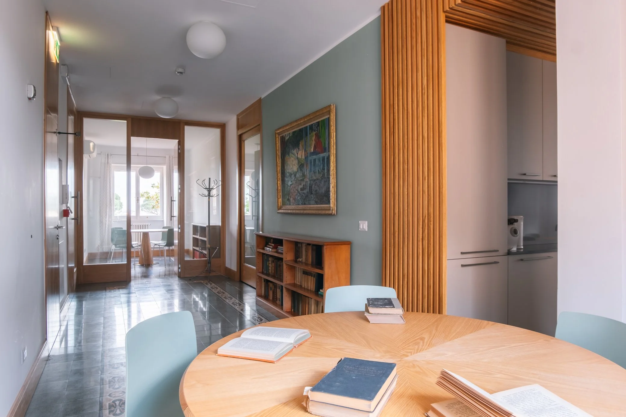 Interno di una casa con sala da pranzo e angolo studio, tavolo rotondo con libri aperti, sedie azzurre e pareti con decorazioni in legno e quadri. Sullo sfondo, zona giorno con finestra grande.