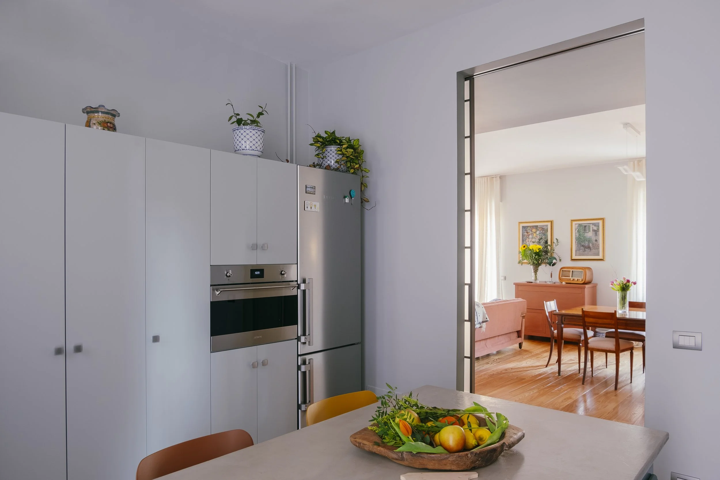 Interno di una casa con cucina e soggiorno. In primo piano, un tavolo con un piatto di frutta e fiori. Sullo sfondo, sono visibili delle pareti decorate con quadri e un mobile con una radio.