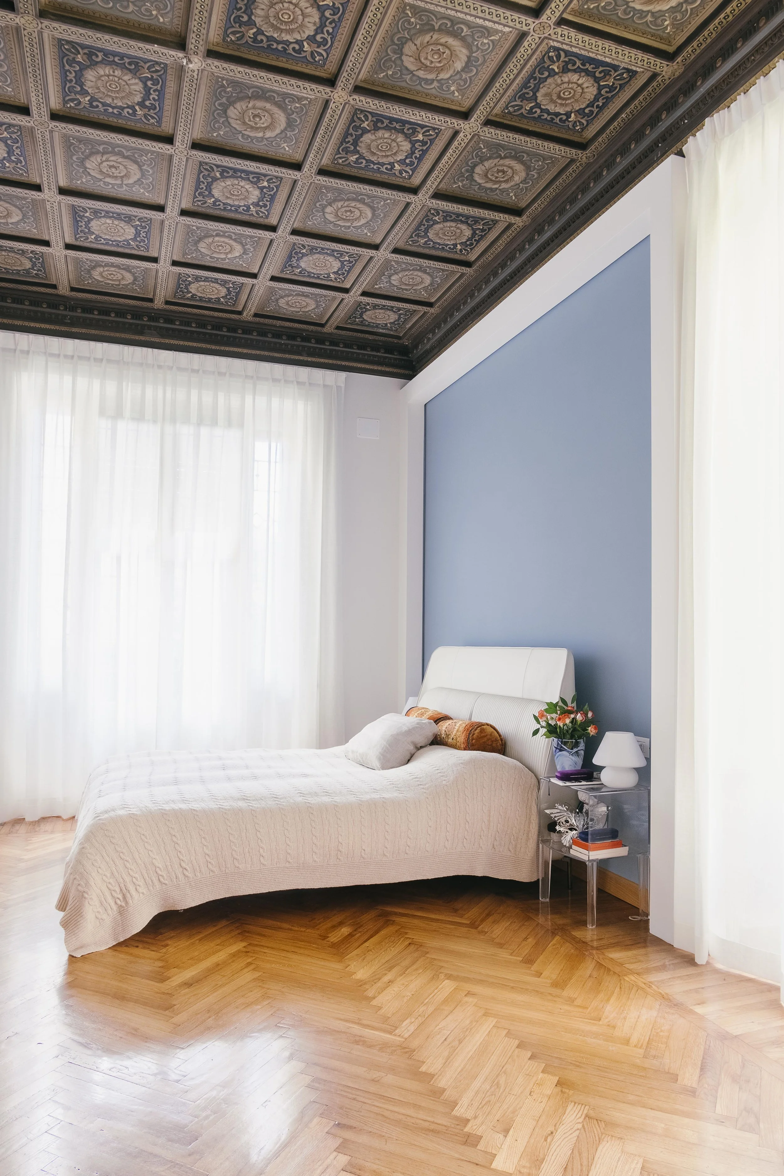 Camera da letto con letto bianco, cuscini, un vaso di fiori, una lampada e una mensola con libri. Pareti bianca e blu, tende bianche e pavimento in parquet di legno.