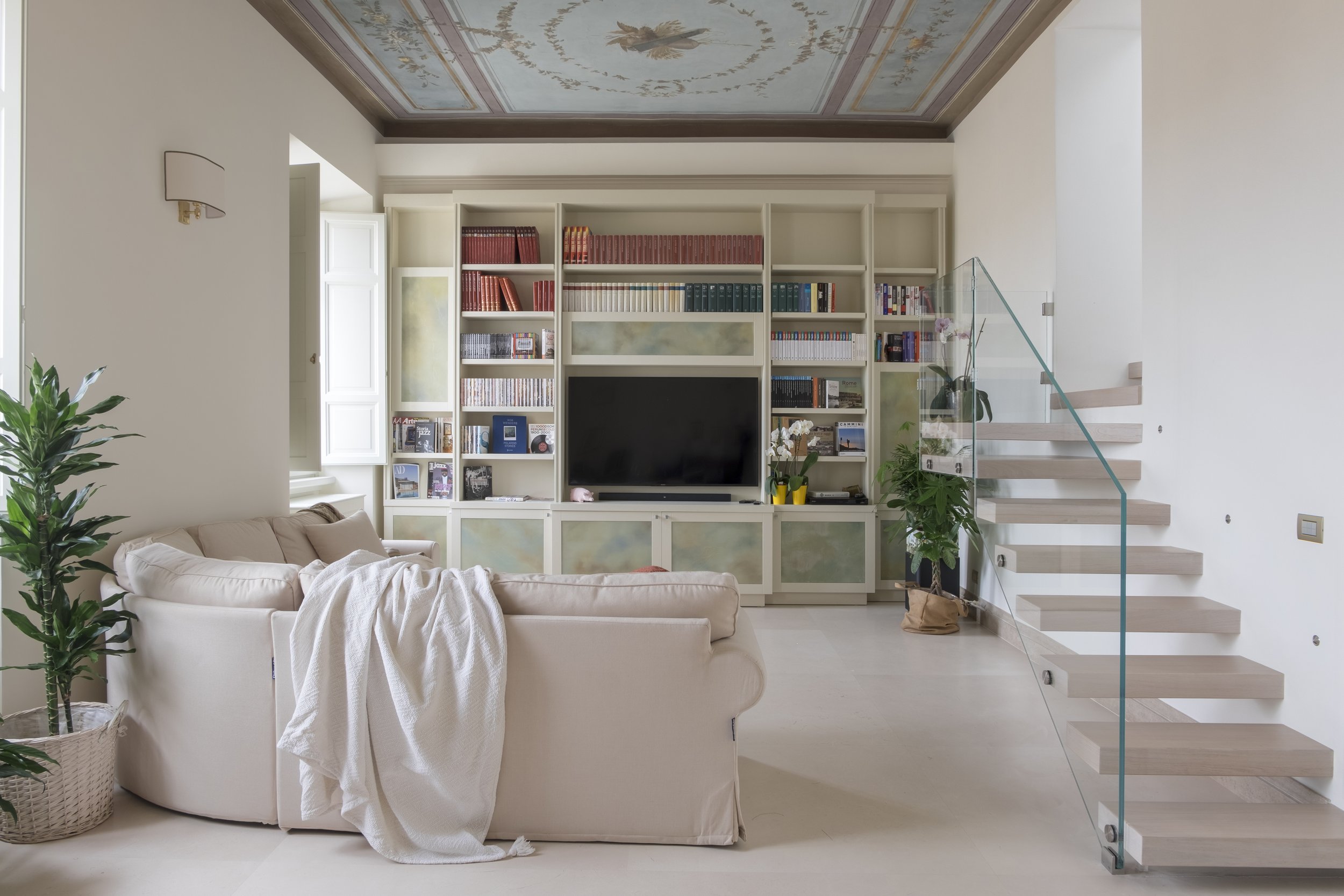 Interior moderno con parete con scaffali pieni di libri, televisore e divano in tessuto chiaro, pianta verde in vaso, soffitto con affresco e scalinata in legno con ringhiera in vetro.