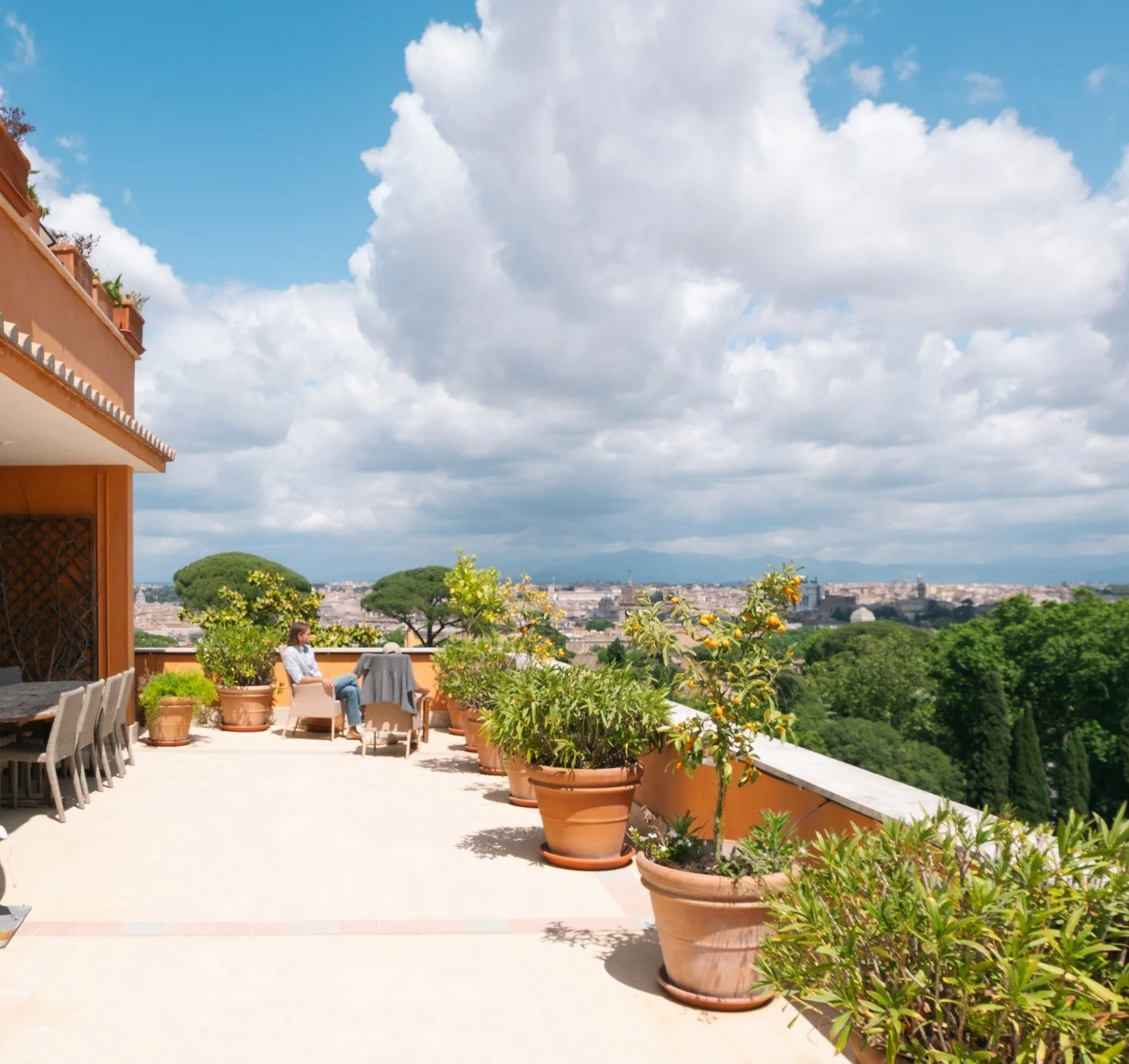 Terrazza con piante in vaso, sedie e tavolo, vista sulla città e sul verde, cielo con nuvole