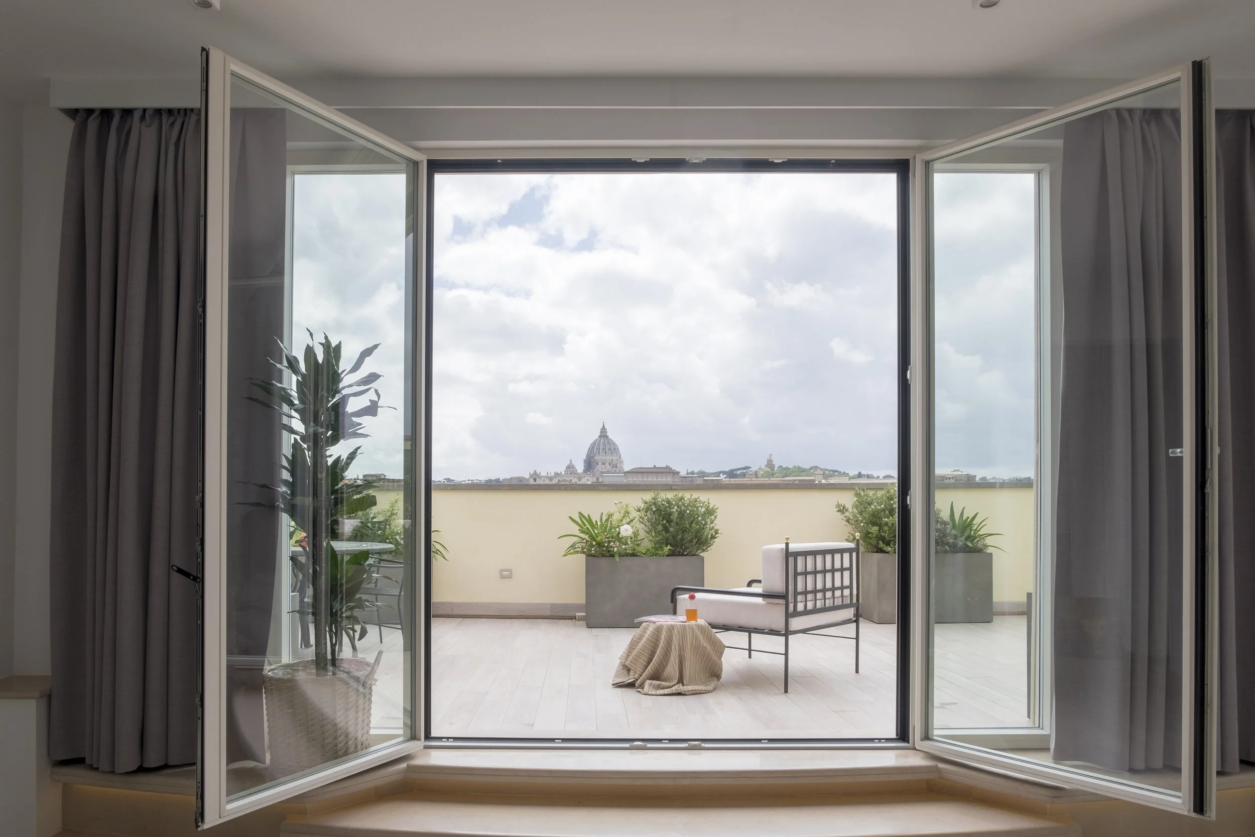 Vista da una finestra aperta su un terrazzo con piante e sedie, con una vista sul Vaticano in lontananza sotto un cielo nuvoloso.