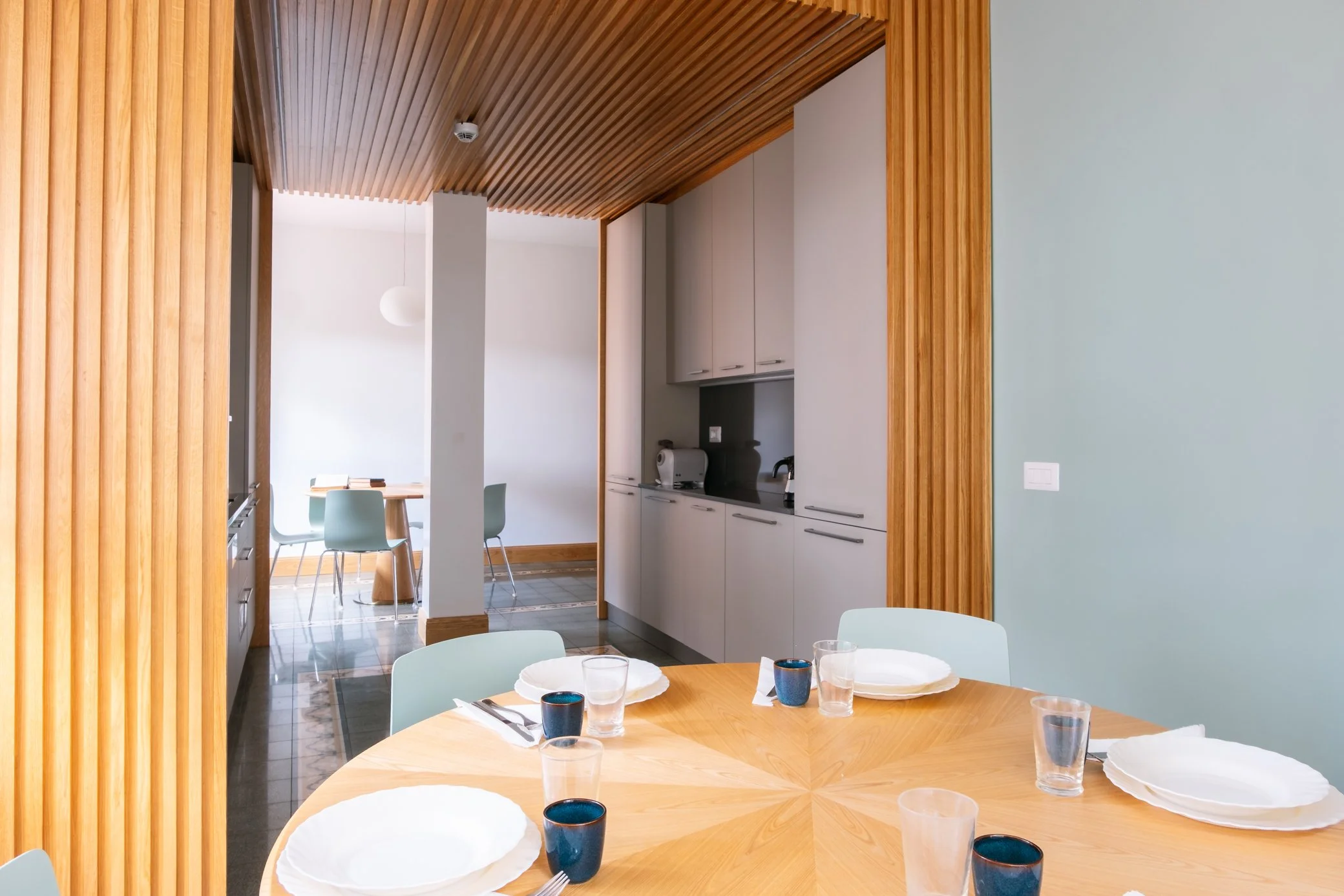 Interno di una sala da pranzo moderna con tavolo rotondo in legno, piatti, bicchieri e tazze, vista verso una cucina con pareti bianche, pensili e un forno, e pareti in bianco e verde, con cucina e spazio per mangiare.