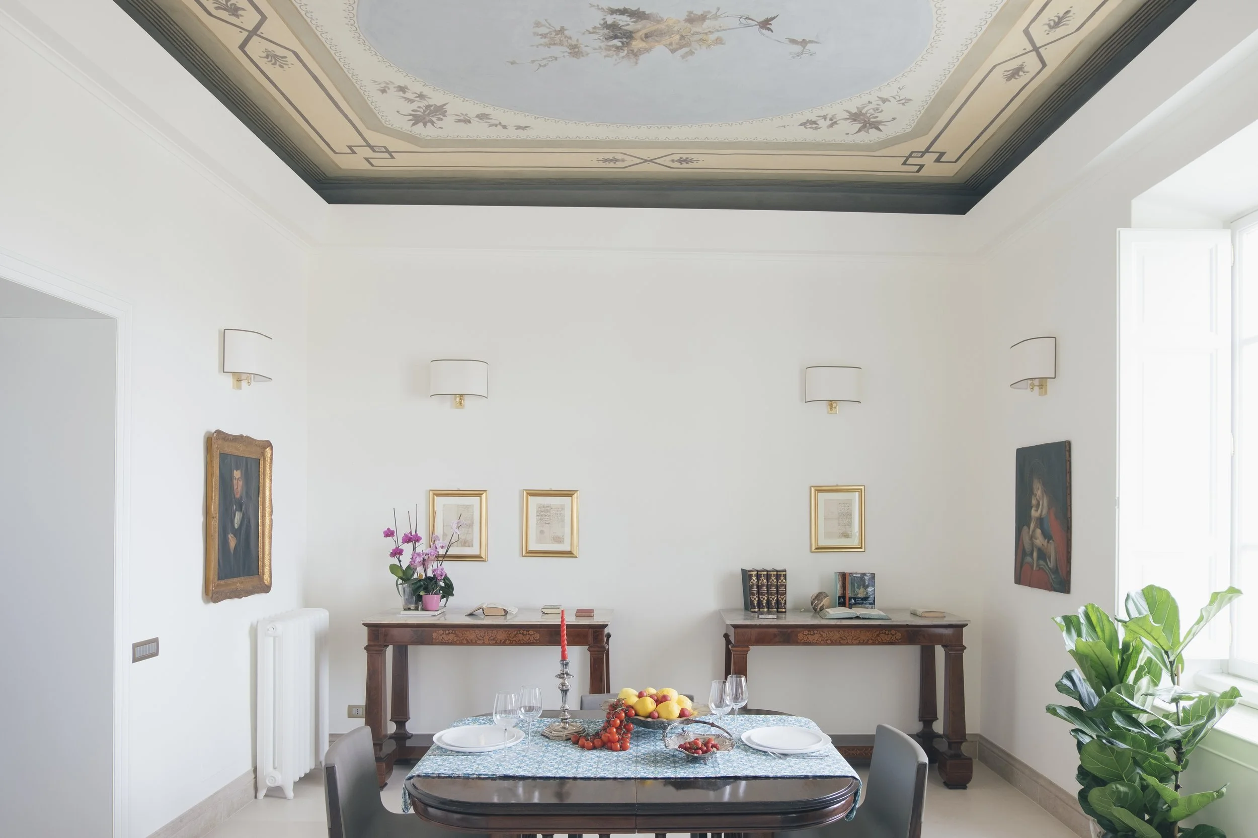 Interno di una sala da pranzo luminosa con pareti bianche, quadri incorniciati, tavolo con tovaglia blu, frutta e candelabro, sedie nere, scaffale con libri, dipinti e piante ornamentali, illuminata da luce naturale.