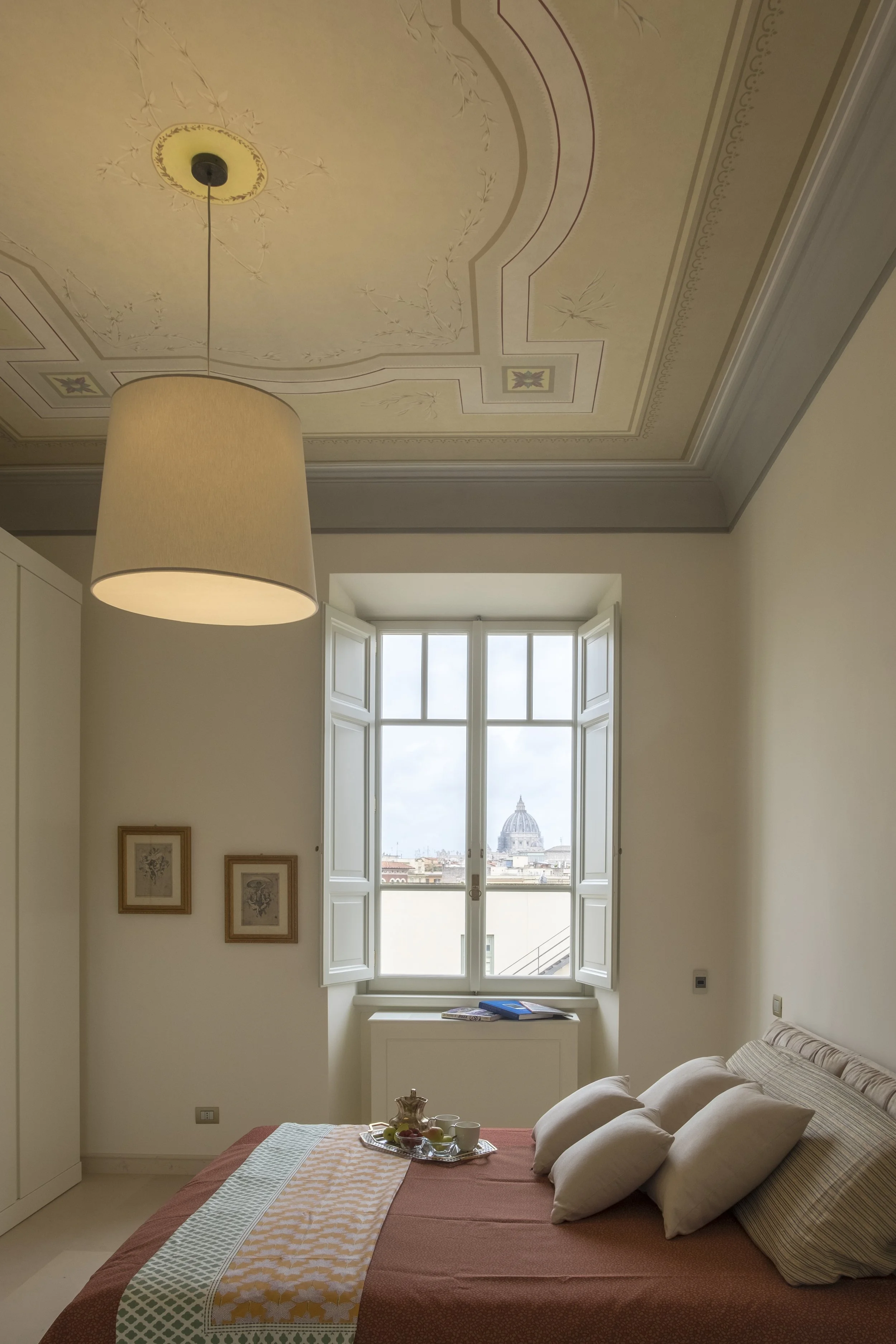 Camera da letto arredata con pareti bianche, letto con coperta color terracotta e diverse cuscini beige e a righe. Sul comodino si trova una teiera e alcune tazze, e c'è una finestra con zanzariera aperta che mostra un panorama con il Duomo di San Pi