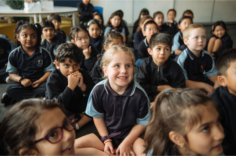 Group of elementary school children sitting on the floor in a classroom, attentively listening and smiling, wearing dark uniforms with light blue accents.