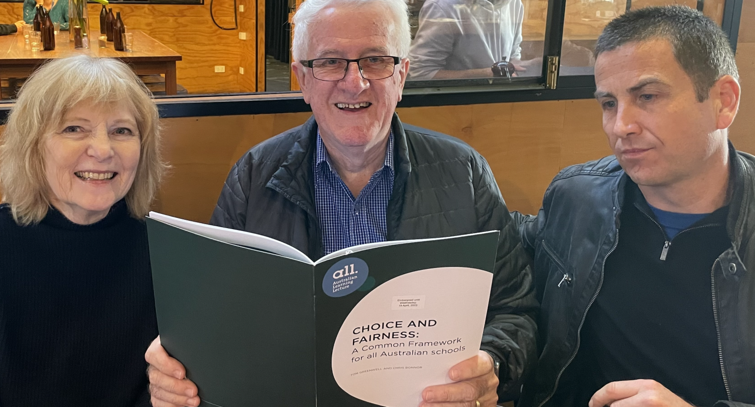 Three people sitting at a table, with a woman on the left, a man in the middle holding a book titled 'Choice and Fairness', and a man on the right, smiling and looking at the book, in a casual indoor setting.