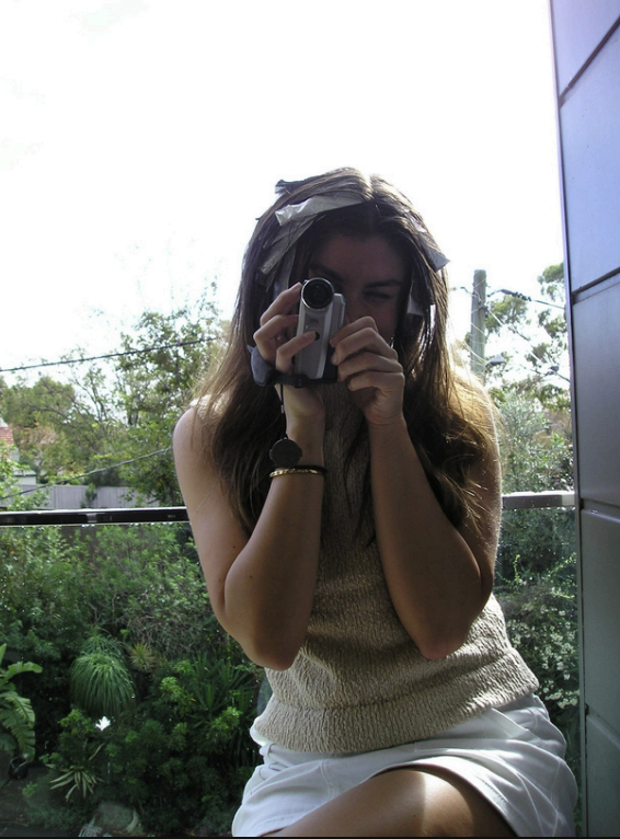 Young woman taking a selfie with a digital camera outside on a balcony, with greenery and trees in the background.